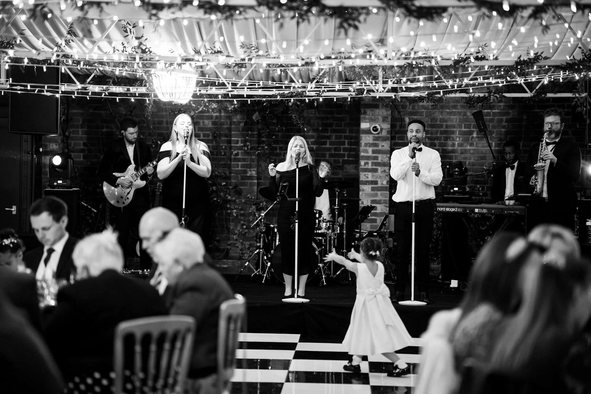 A band performs on a lit stage beneath fairy lights while diners sit below; a small child in a dress dances on the checkered floor in front.