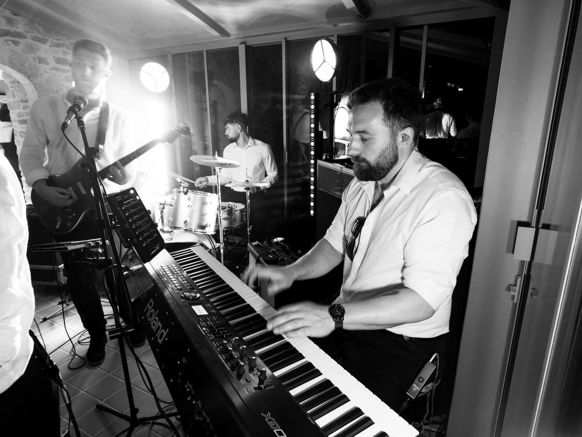 Black-and-white studio photo shows a keyboardist playing in front, with a guitarist and drummer behind him; the intimate band session feels lively.