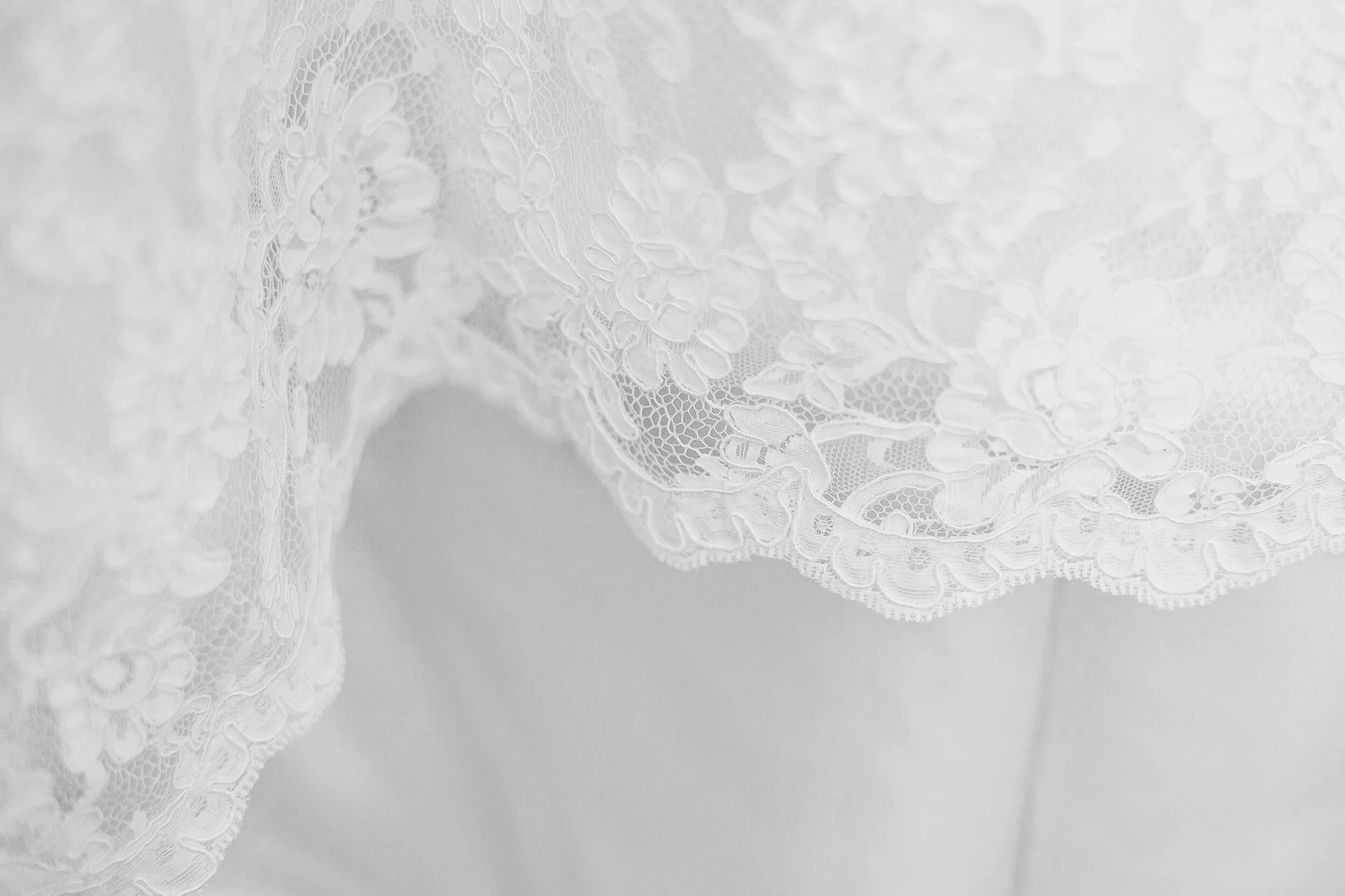 Close-up of delicate lace fabric with floral patterns and scalloped edges, softly draped—likely part of a wedding dress, light and intricate.