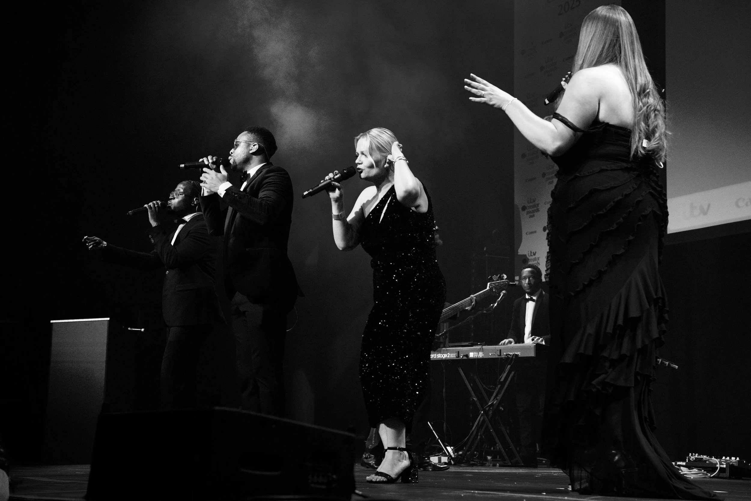 Four singers perform on stage with a live band behind them, holding microphones and singing under stage lights in a formal concert setting.