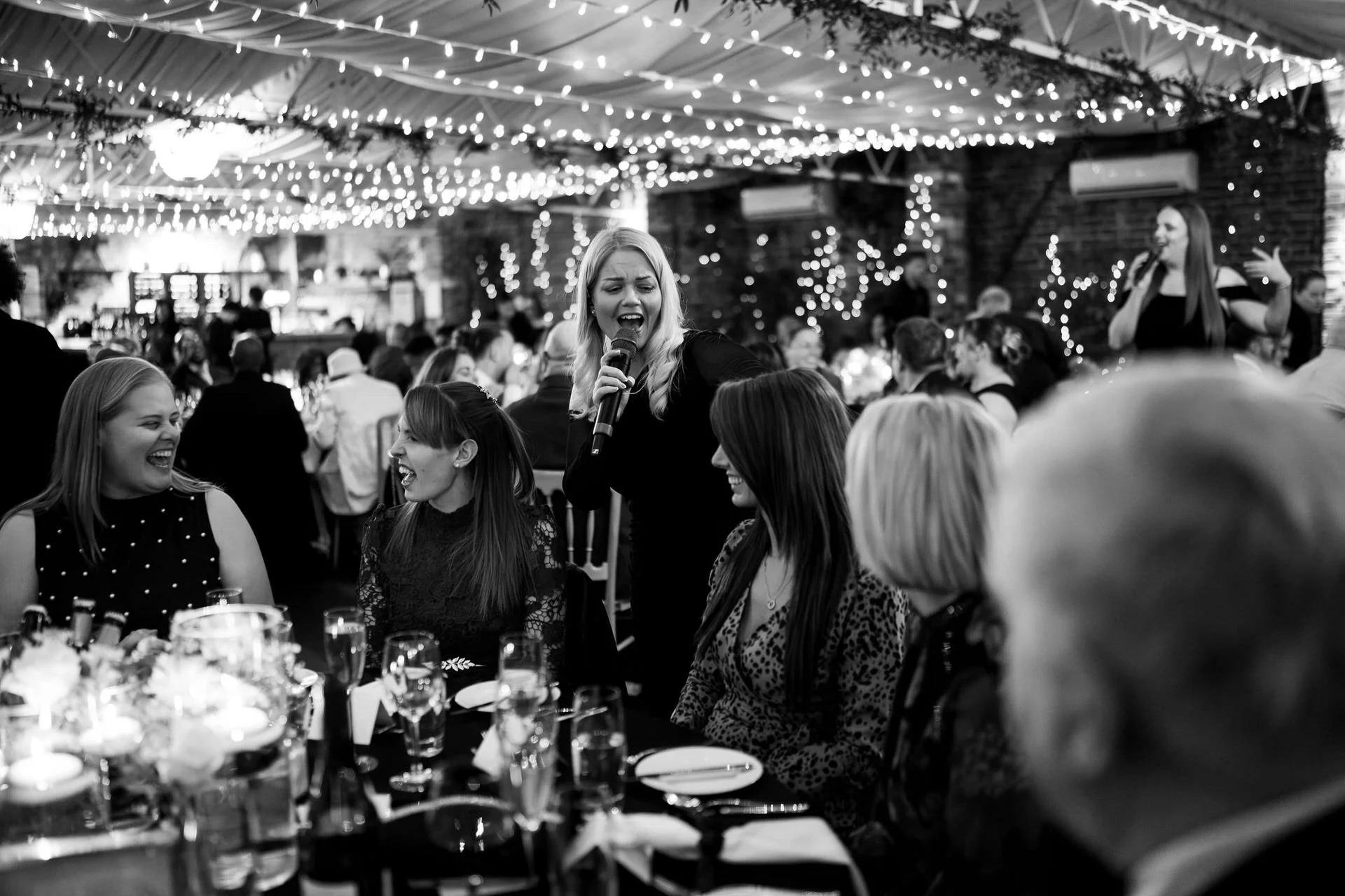 A singer leans into a candlelit table, performing for laughing guests beneath twinkling fairy lights in a crowded, elegant banquet hall.