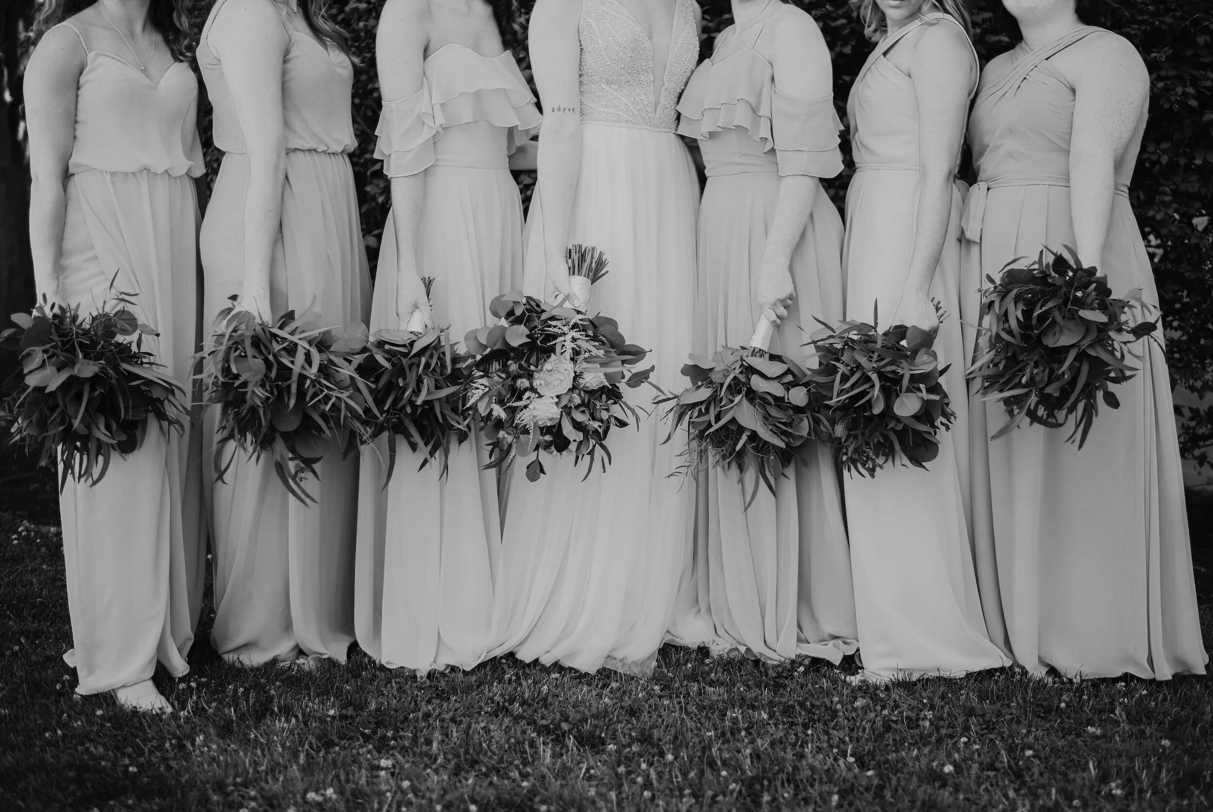 Six bridesmaids in long dresses stand side by side on grass, each holding leafy bouquets; the bride stands among them with a fuller central bouquet.