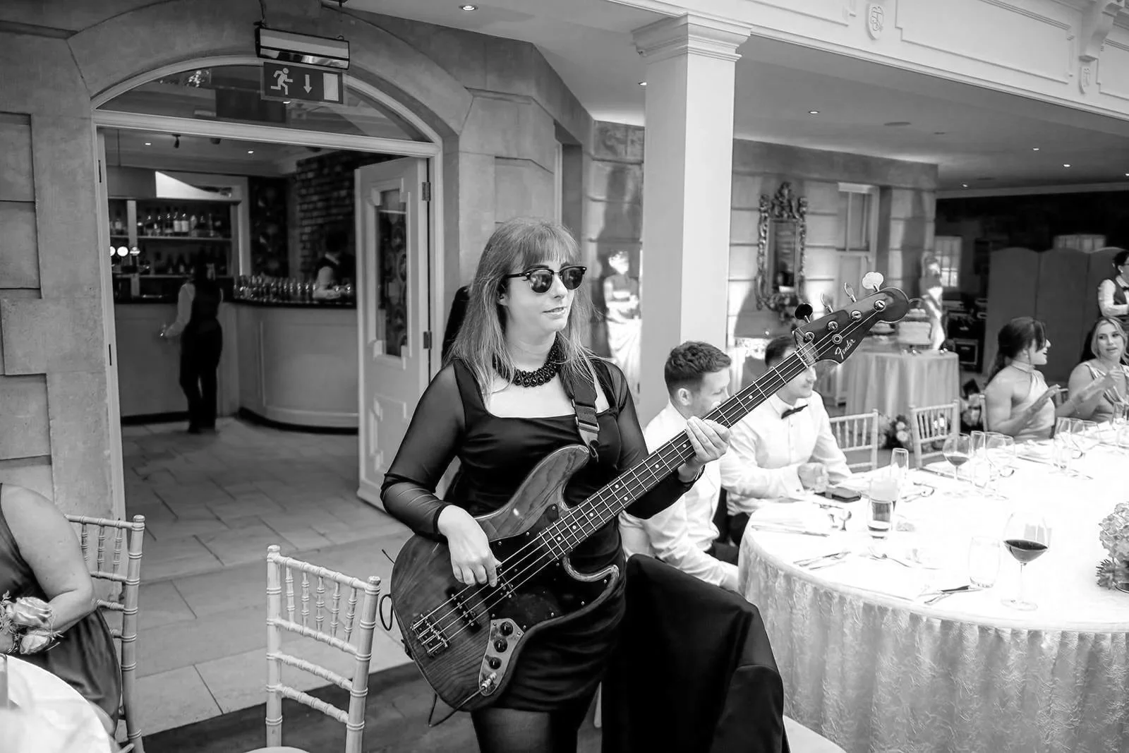 A woman in a black dress and blue sunglasses plays bass guitar among seated wedding guests in a bright, elegant room.