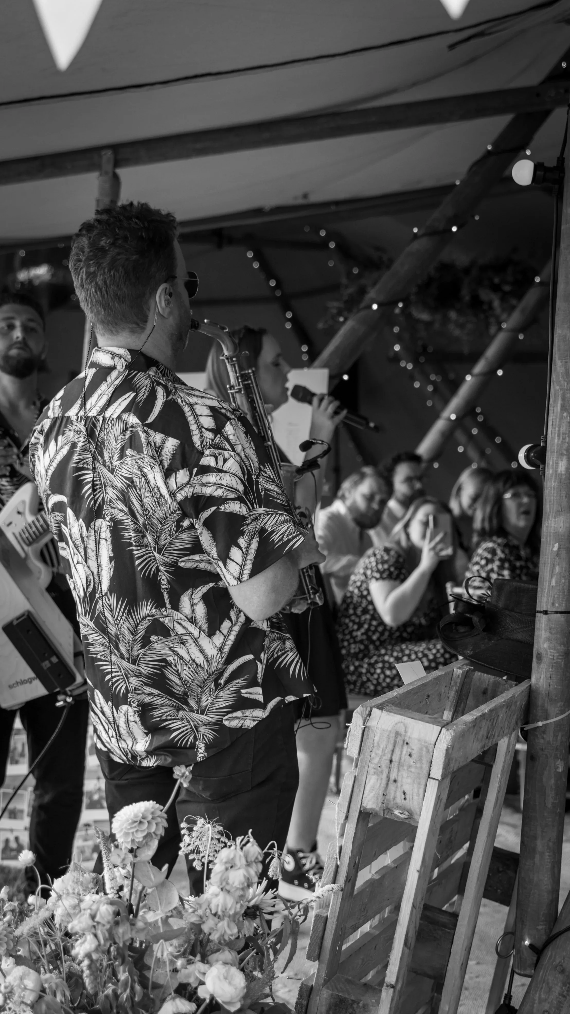 A saxophonist in a patterned shirt plays facing a crowd. A singer performs beside him as guests watch, some filming, under soft string lights.