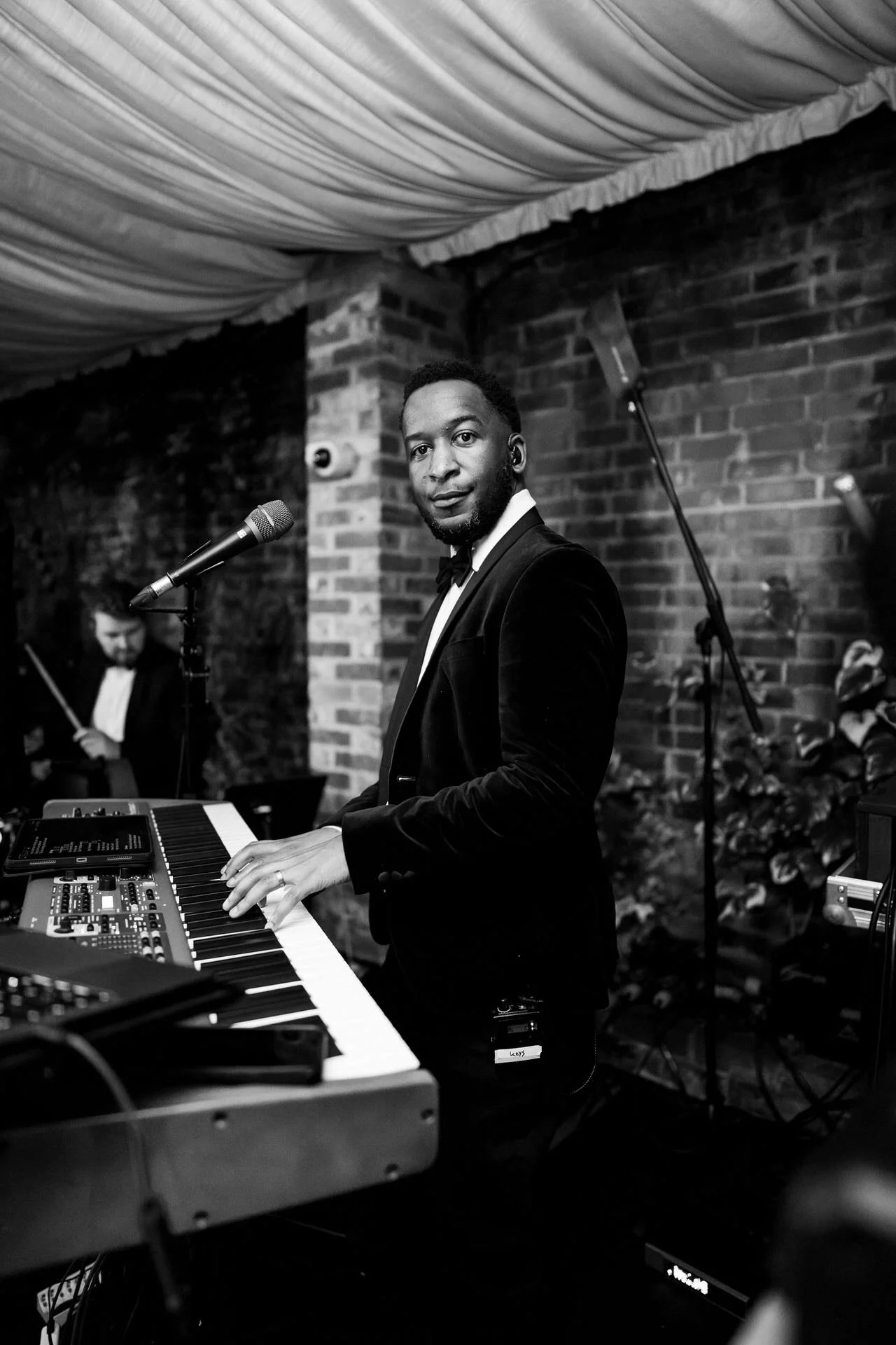 A black-and-white photo of a tuxedoed musician at a keyboard, glancing toward the camera, with microphones and brick walls behind him.
