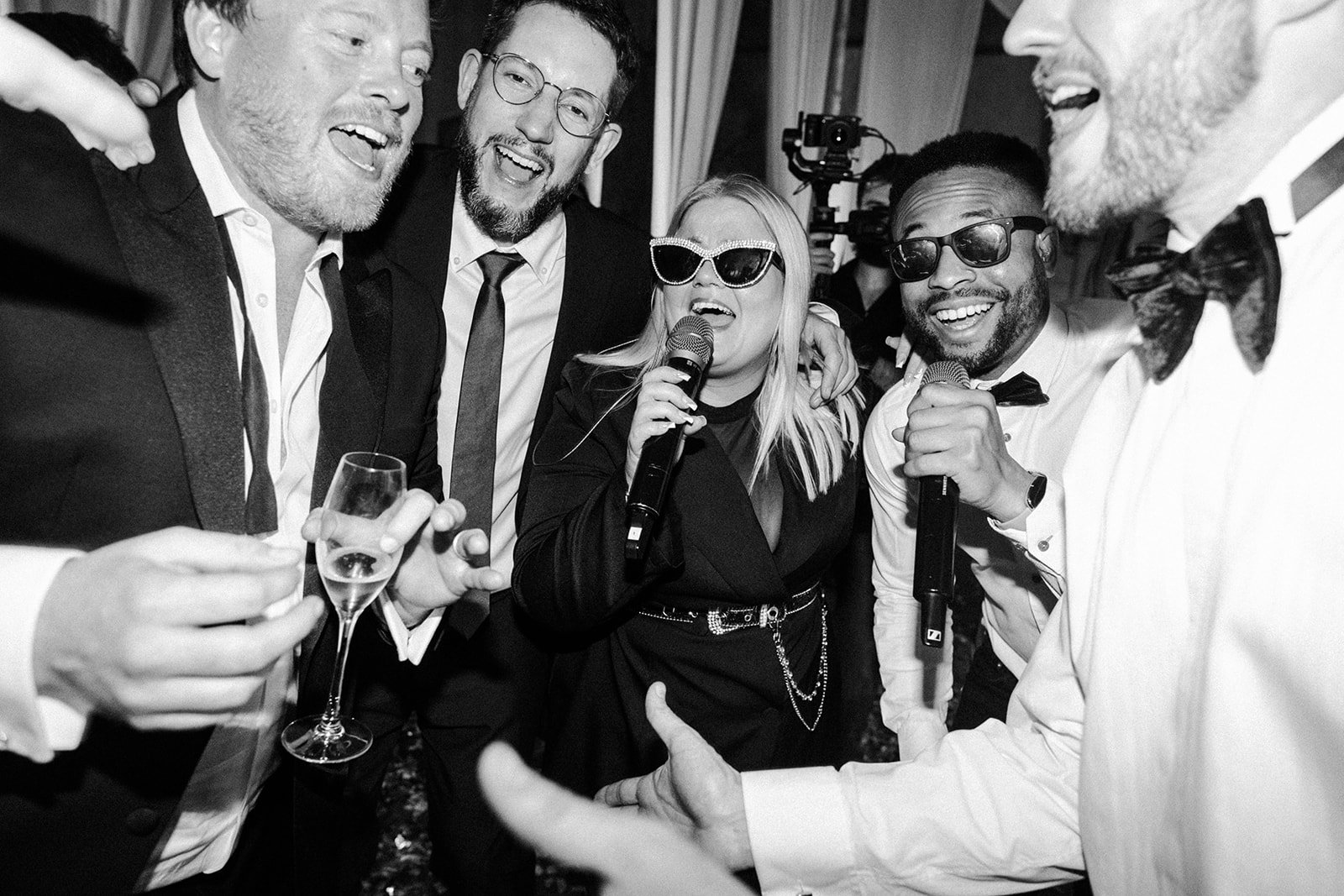 Five well-dressed adults crowd together, laughing and singing into microphones; one holds a champagne flute. It feels loud, joyful, and celebratory.
