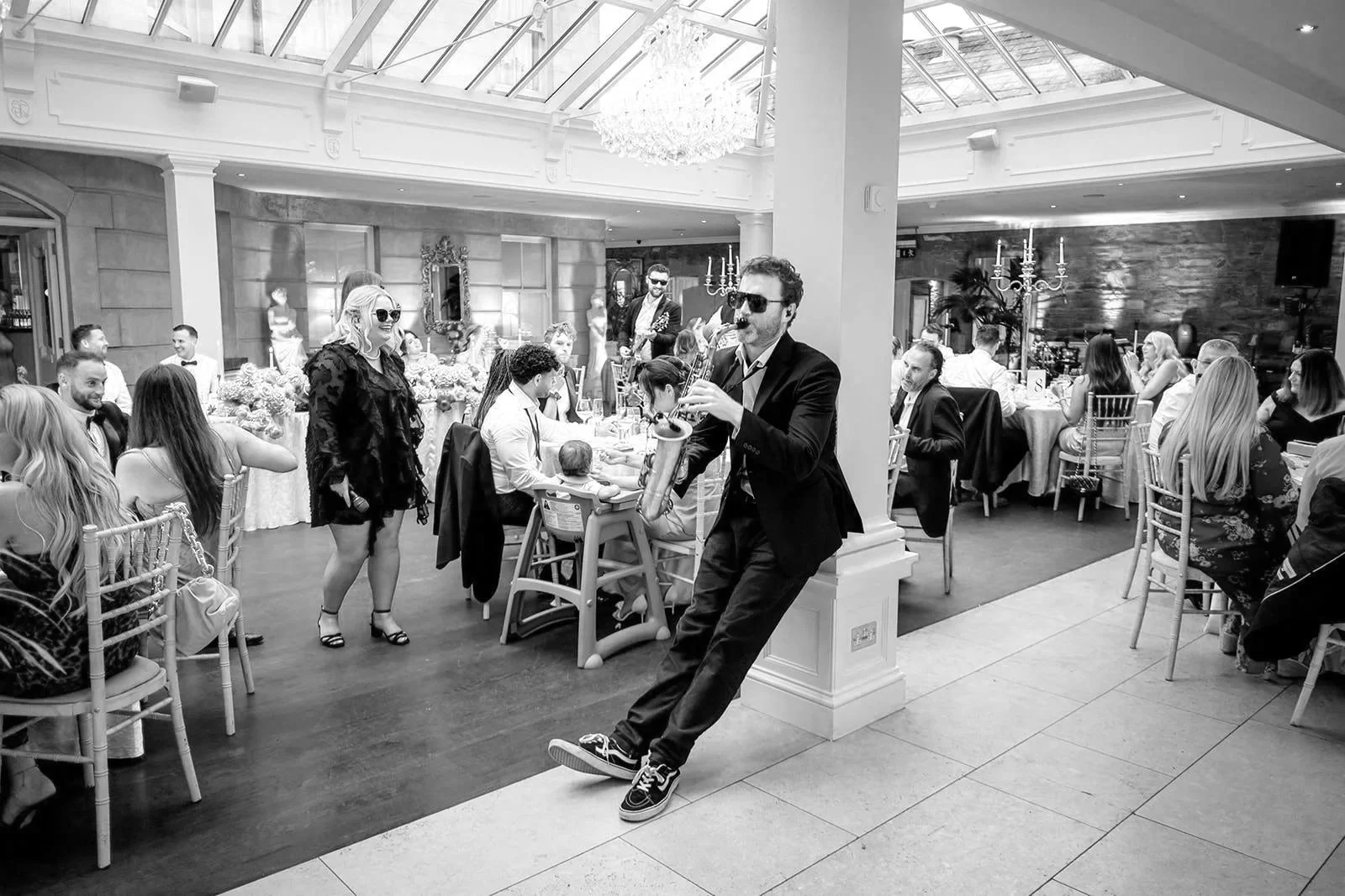 At Tankardstown House, Ireland, a NeoRoots sax player in sunglasses leans and plays among wedding guests under a glass roof and chandelier.