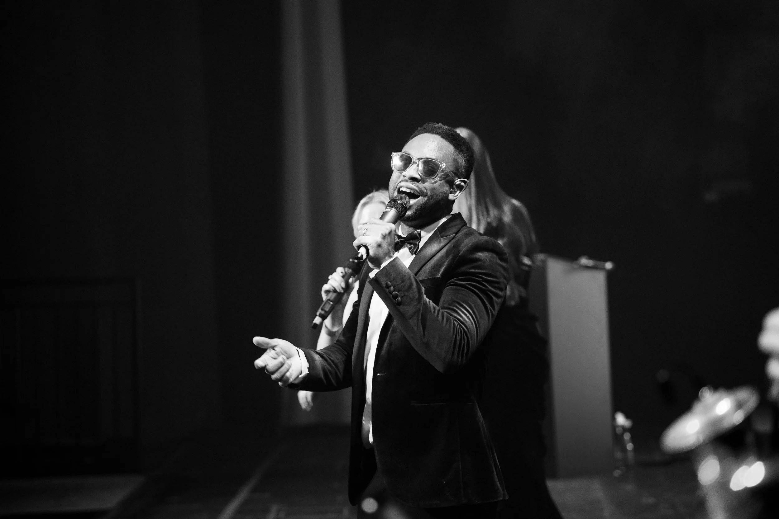 NeoRoots vocalist sings into a microphone on stage in formal attire during a live performance at a London corporate awards event.