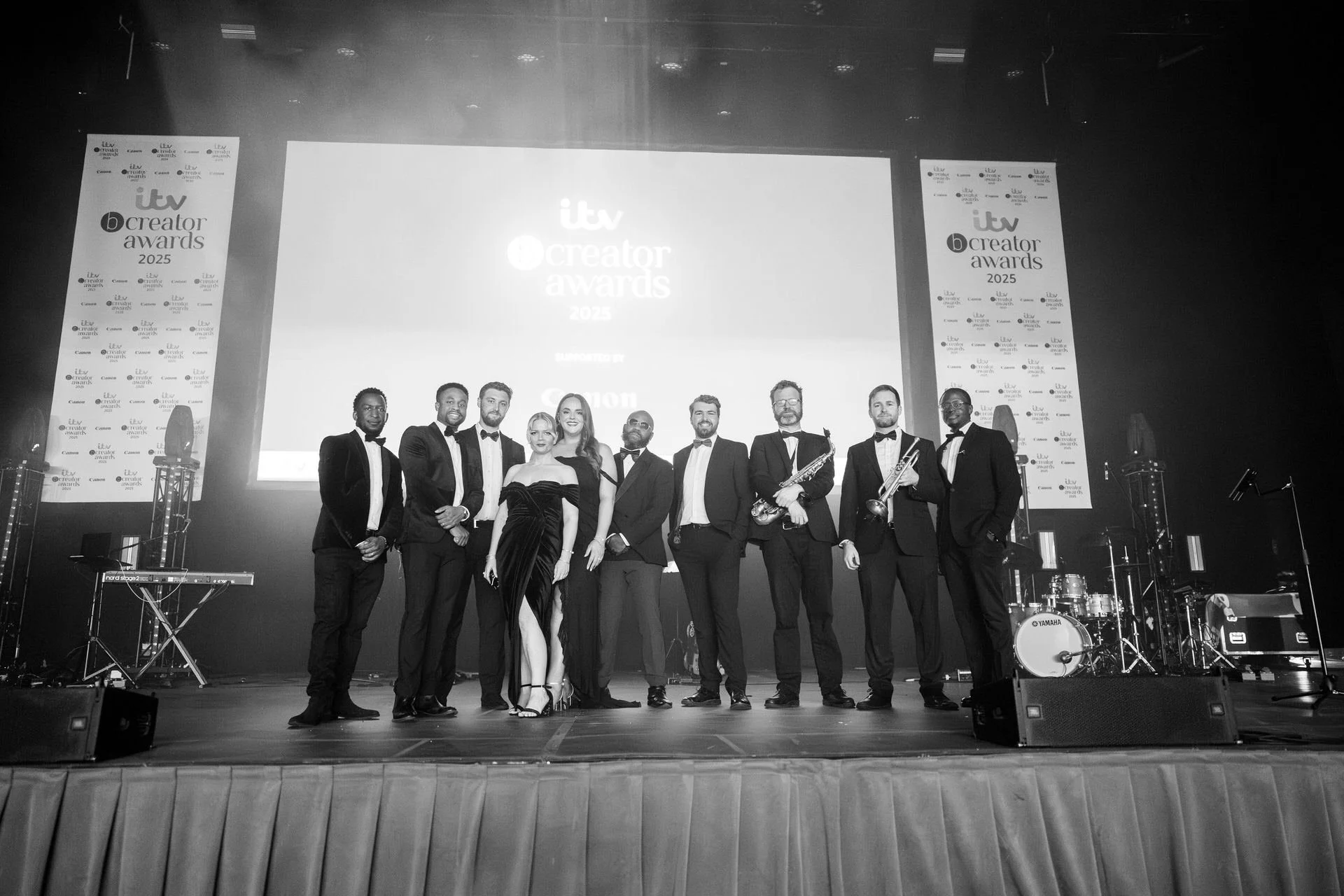A large band in tuxedos poses on a stage beneath a big awards screen, instruments beside them, after a formal performance.