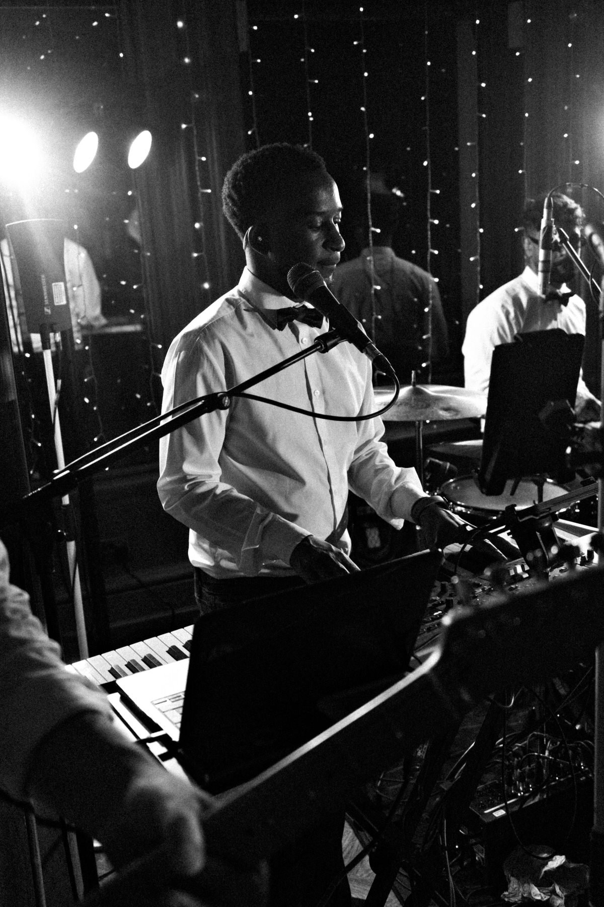 Black-and-white photo of a focused musician playing keyboard and singing into a microphone, surrounded by bandmates, cables, and stage lights.