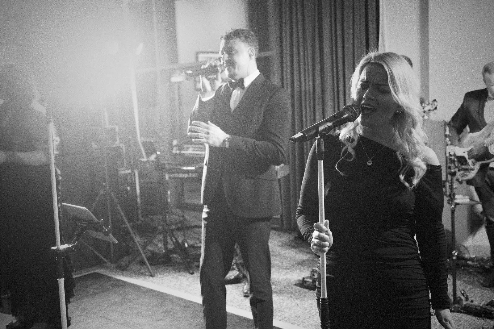 Black-and-white image of two singers performing: a woman gripping a mic stand in front, a man singing behind her, band equipment around them.