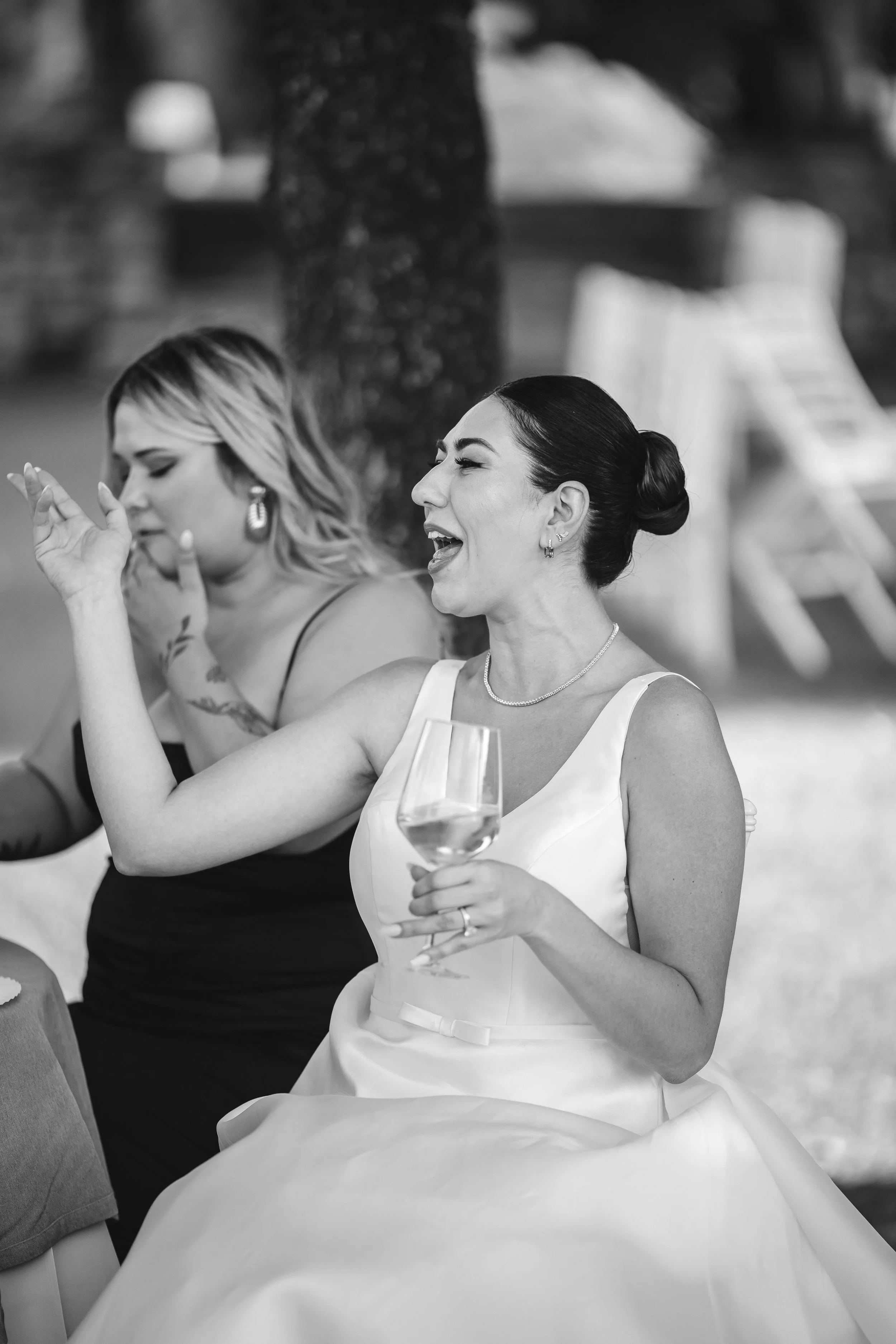 A bride in a flowing gown laughs and gestures mid-toast, holding a wine glass, while a friend beside her wipes away tears at an emotional wedding moment.
