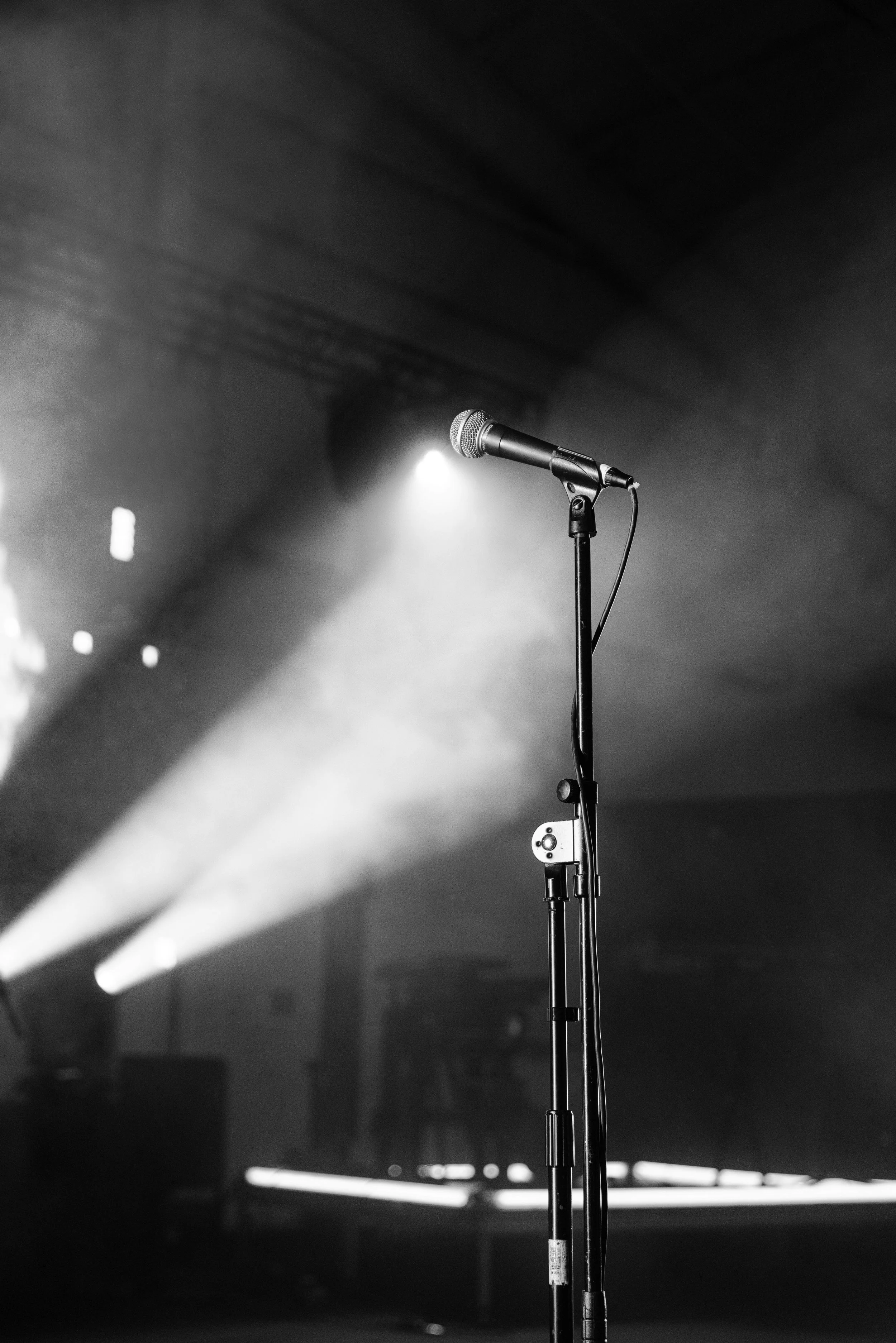 A lone microphone on a stand glows under bright spotlights, with pale beams cutting through smoky air and fading into darkness behind it.