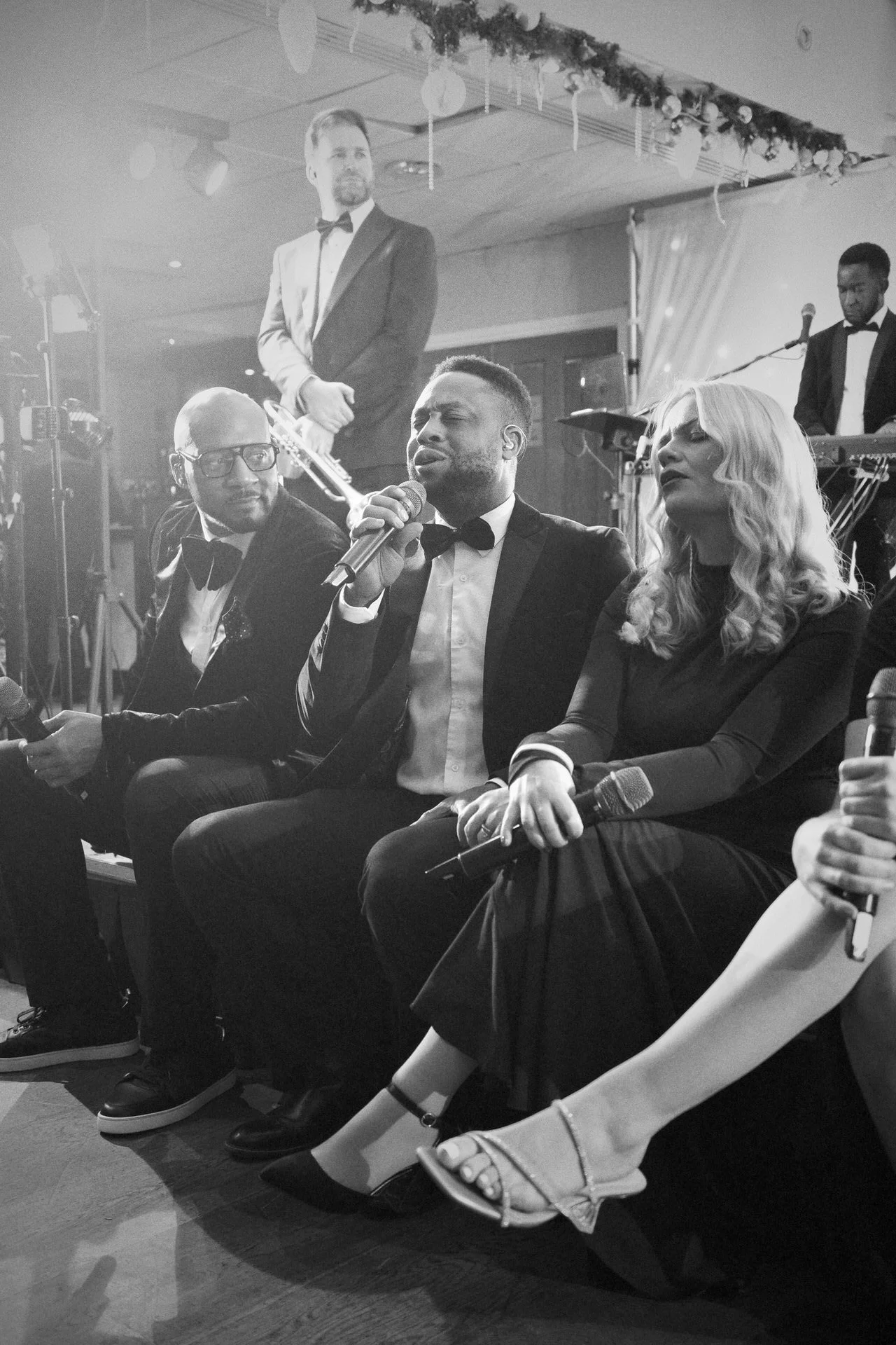 Four formally dressed singers sit at the stage edge holding microphones, performing while other band members play behind them under soft lights.