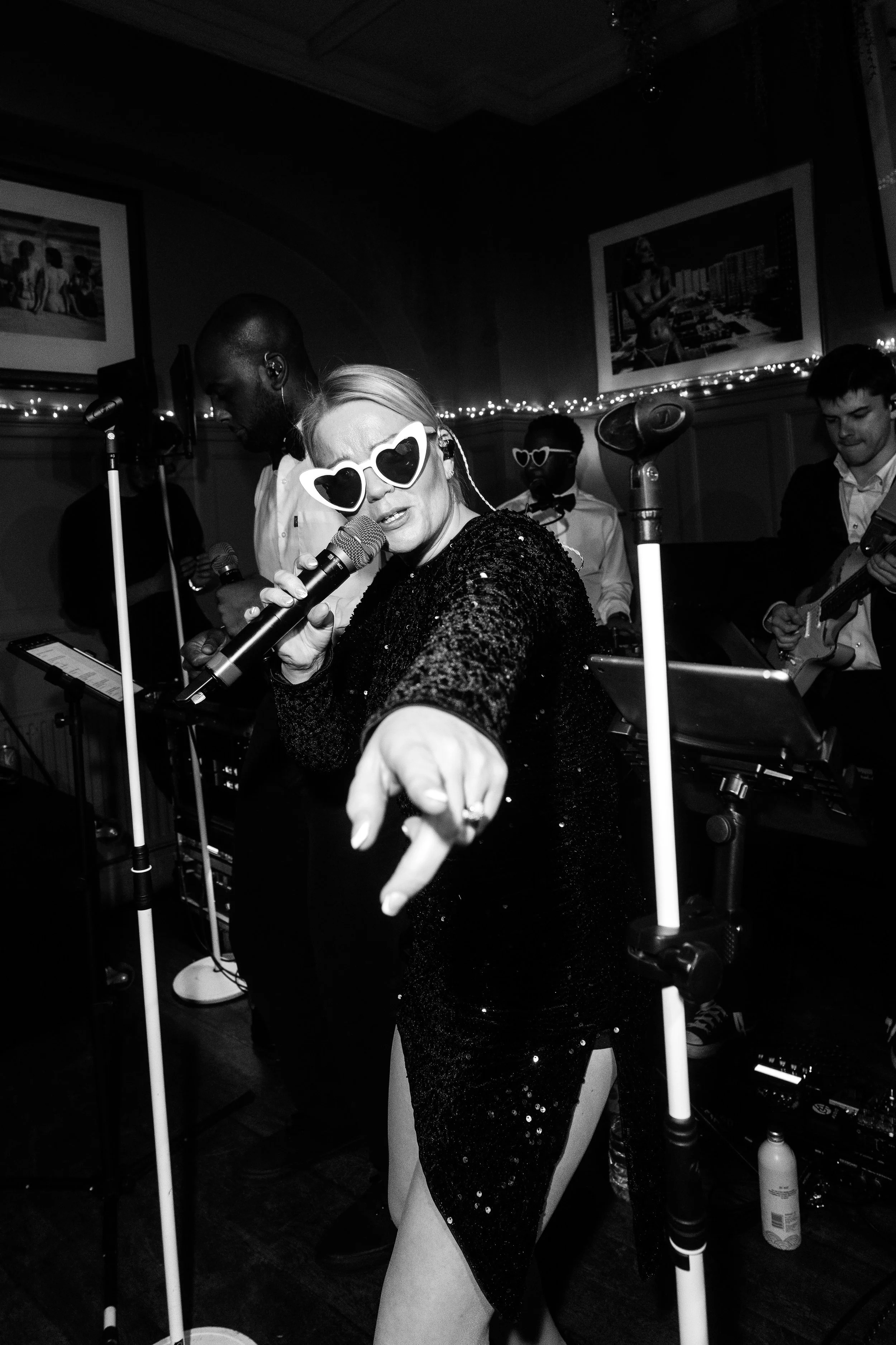 Black-and-white shot of a singer in heart-shaped sunglasses pointing toward the crowd, mic in hand, band playing behind her.