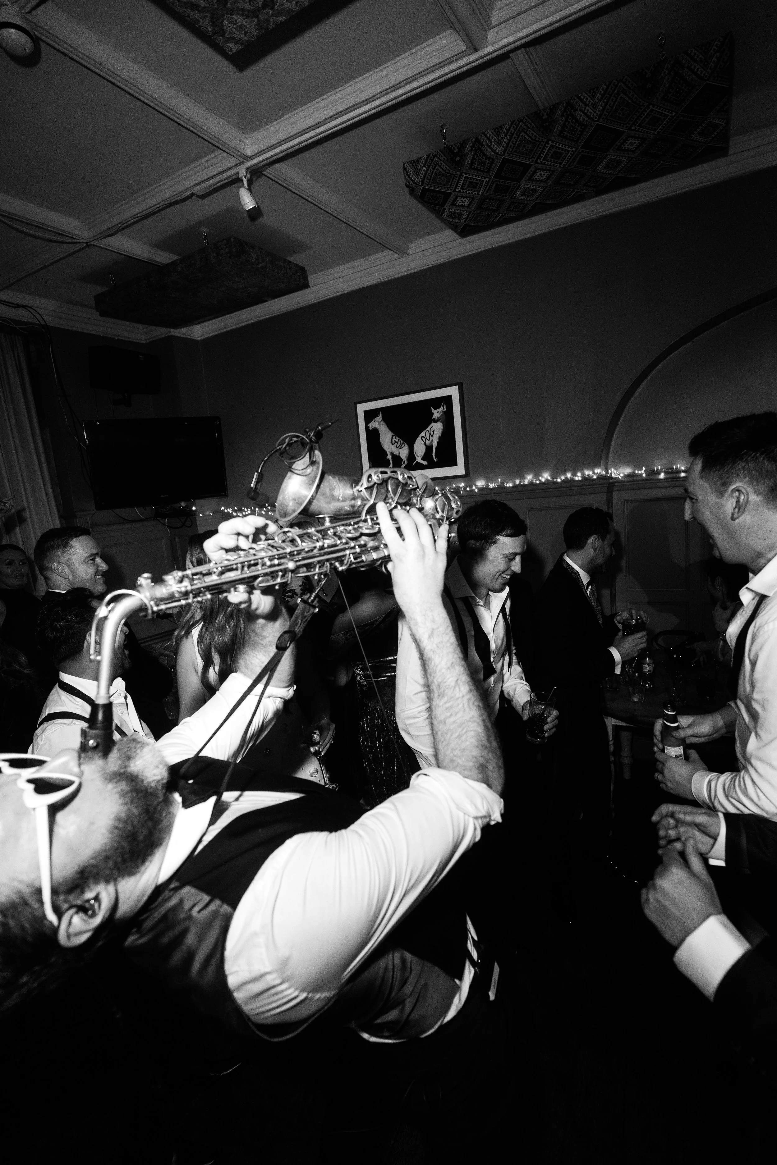 Black-and-white scene of a sax player leaning back and playing above a lively crowd in a dim room with fairy lights.