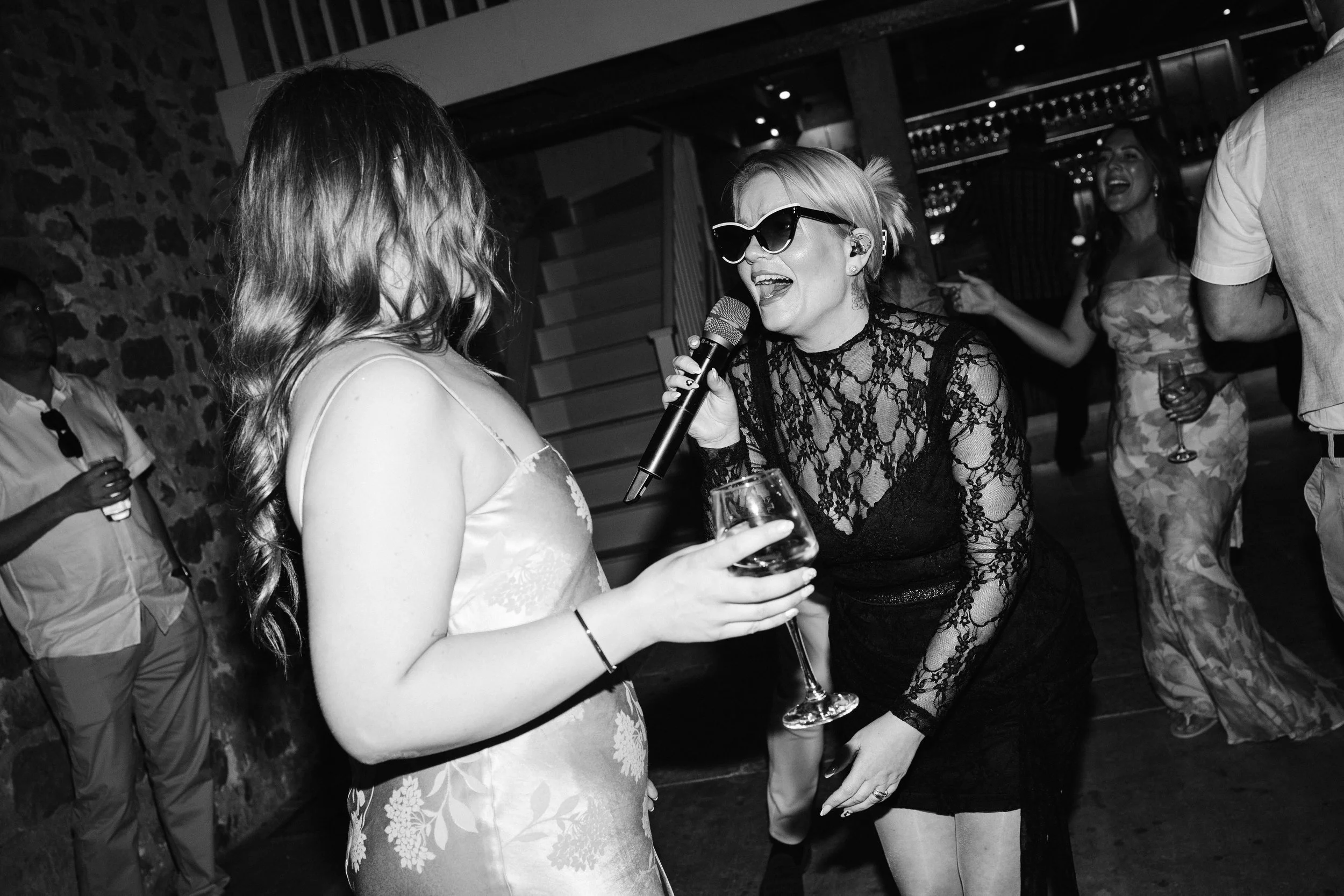 Black-and-white scene of a singer in lace and sunglasses leaning in, singing to a smiling woman holding a wine glass on the dance floor.