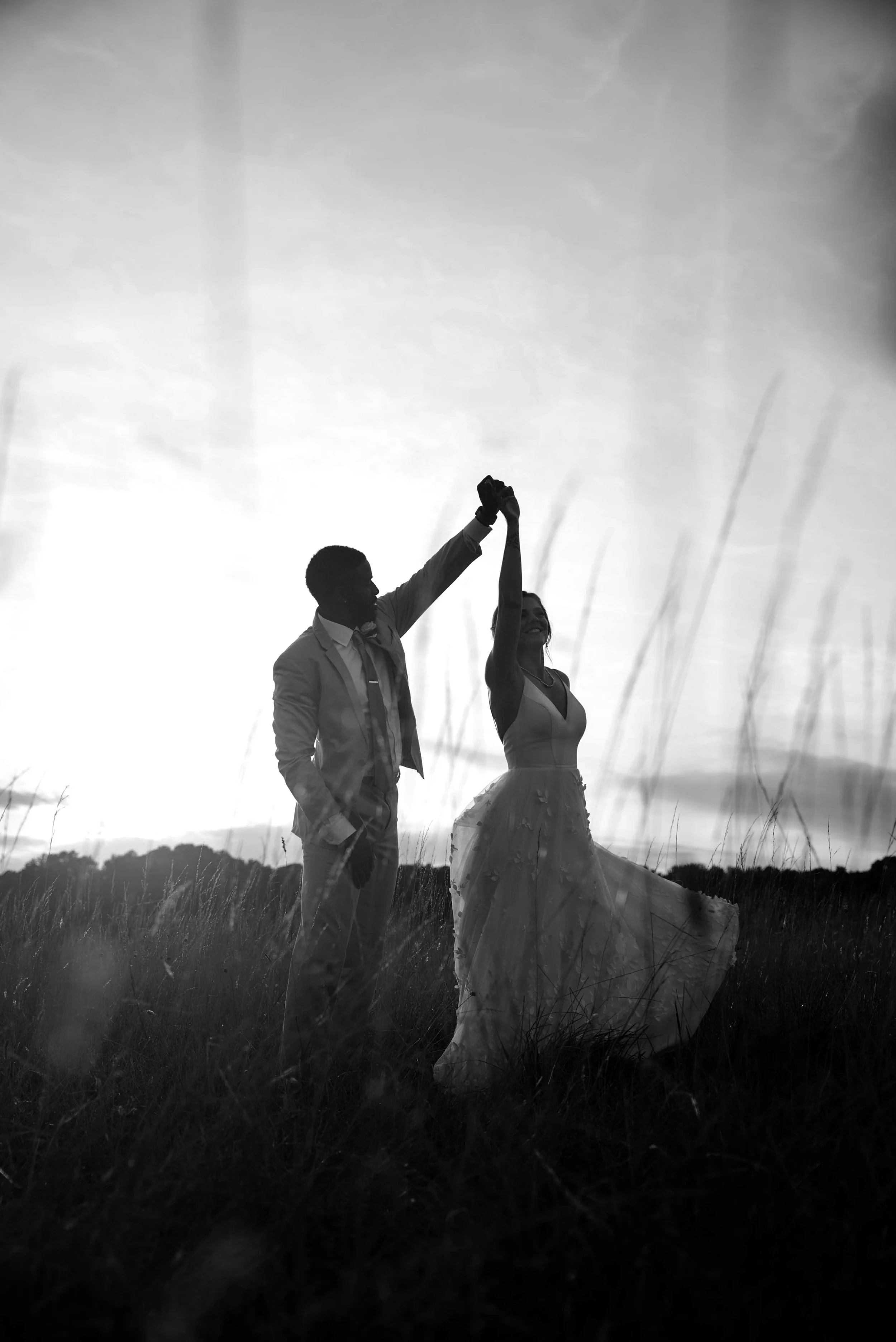 A couple dances in tall grass at sunset, hands raised, silhouetted against the sky as the bride’s dress lifts in the breeze.