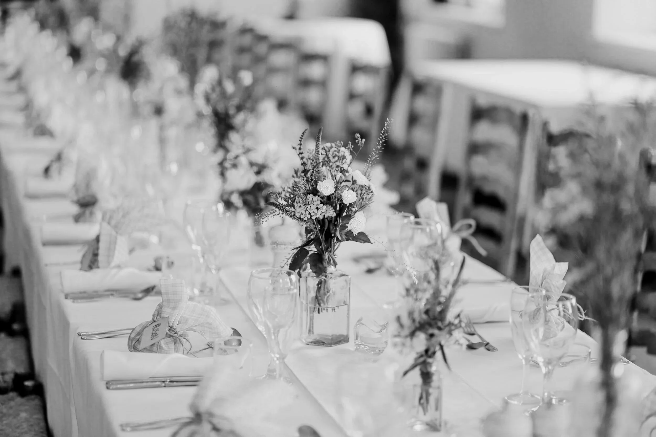 A long banquet table is neatly set with plates, glasses, folded napkins, and small jars of wildflowers, creating a calm, elegant wedding setting.
