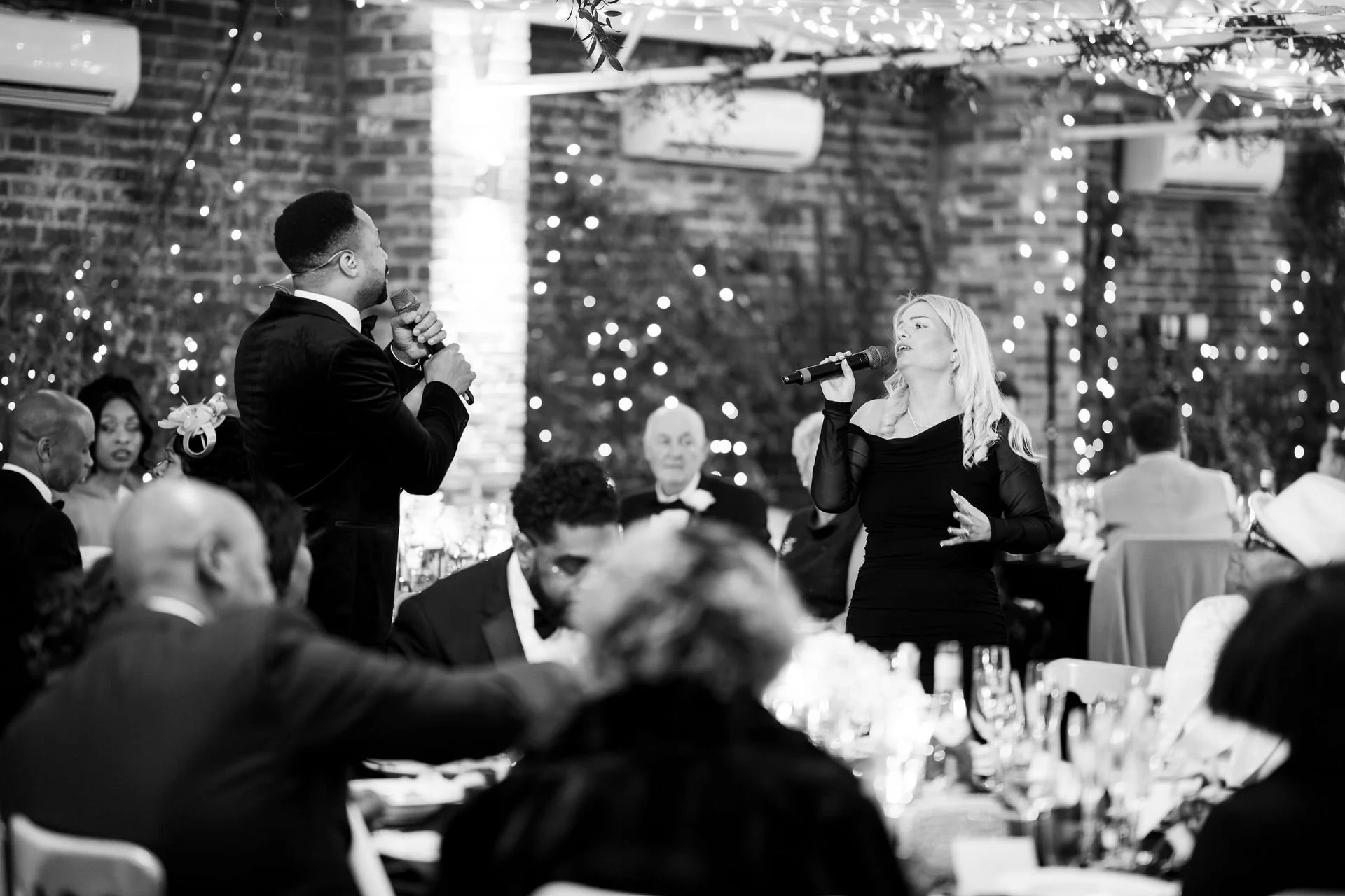Two singers face each other among seated guests, performing at candlelit tables beneath twinkling lights and brick walls during an elegant indoor event.