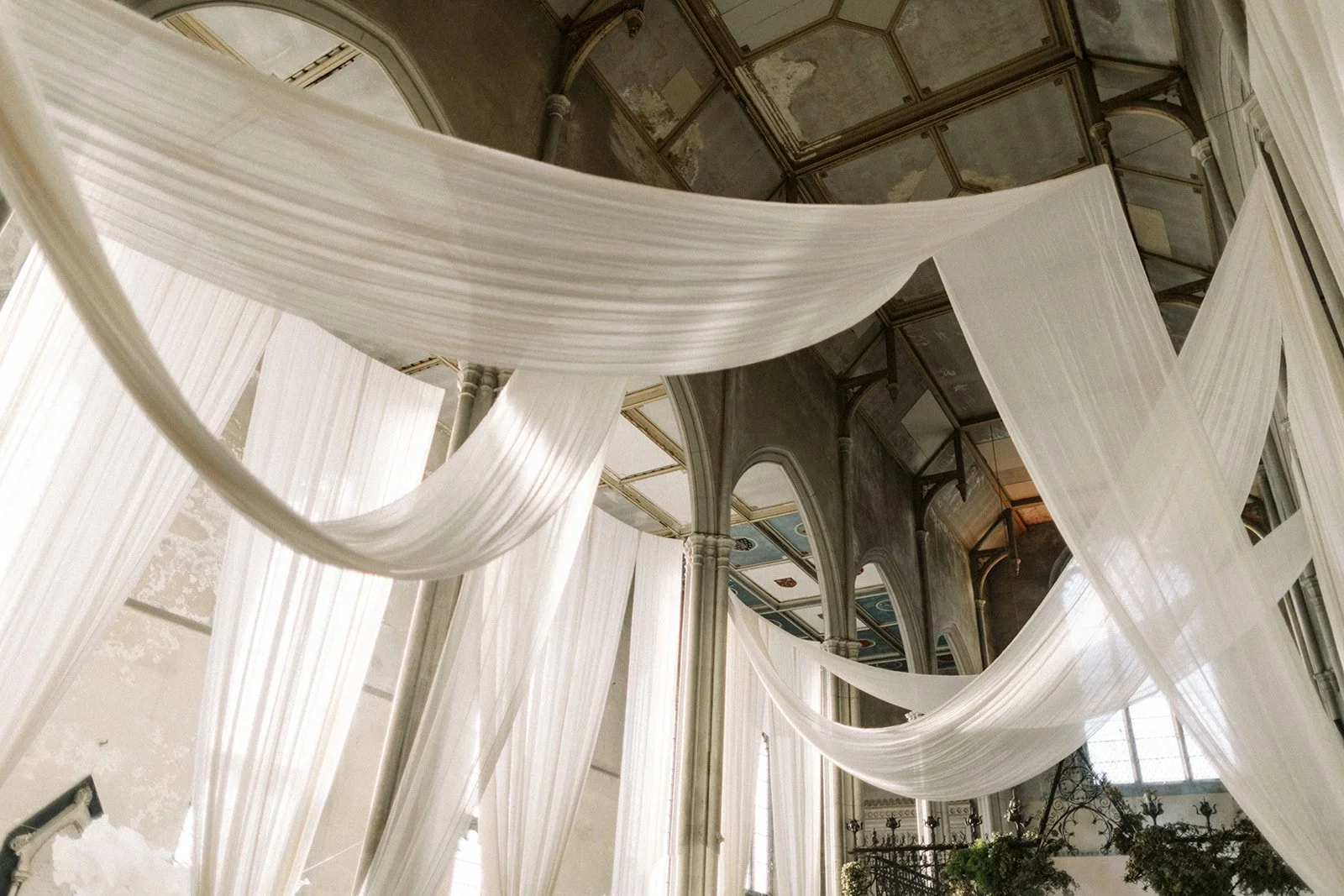Sheer white fabric is draped in soft waves across a high, arched ceiling in a grand hall, creating an airy, romantic atmosphere.