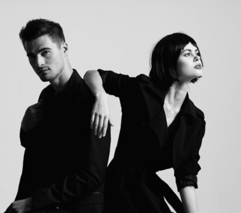 Black and white photo of a young man and woman posing together against a plain background. The man is dressed in a dark suit, looking at the camera, while the woman, with short dark hair and wearing a dark dress, is looking off to the side with her arm resting on the man's shoulder.