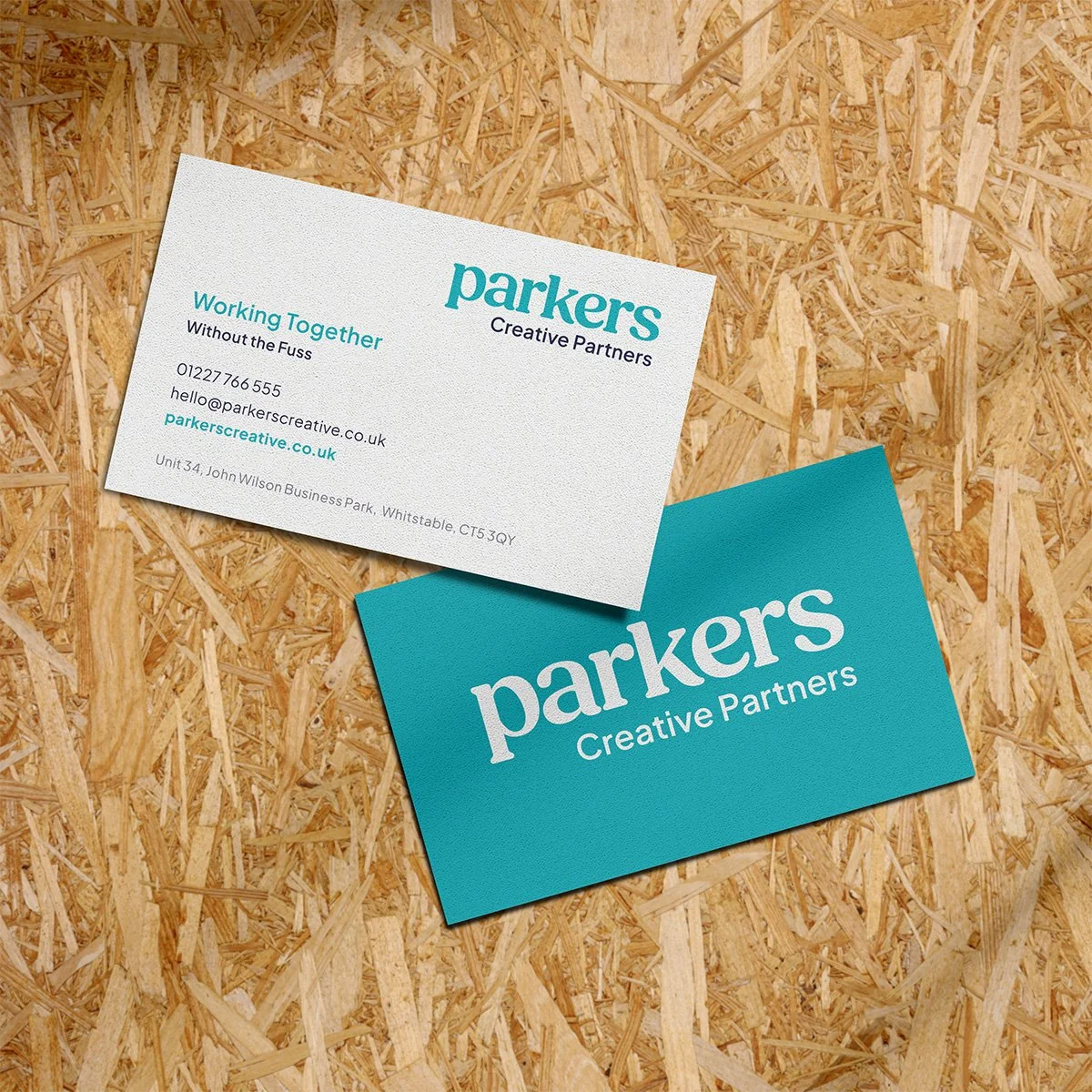 A business card for Parkers Creative Partners placed on a wooden surface. The card displays the logo, contact information, and website in teal and black text.