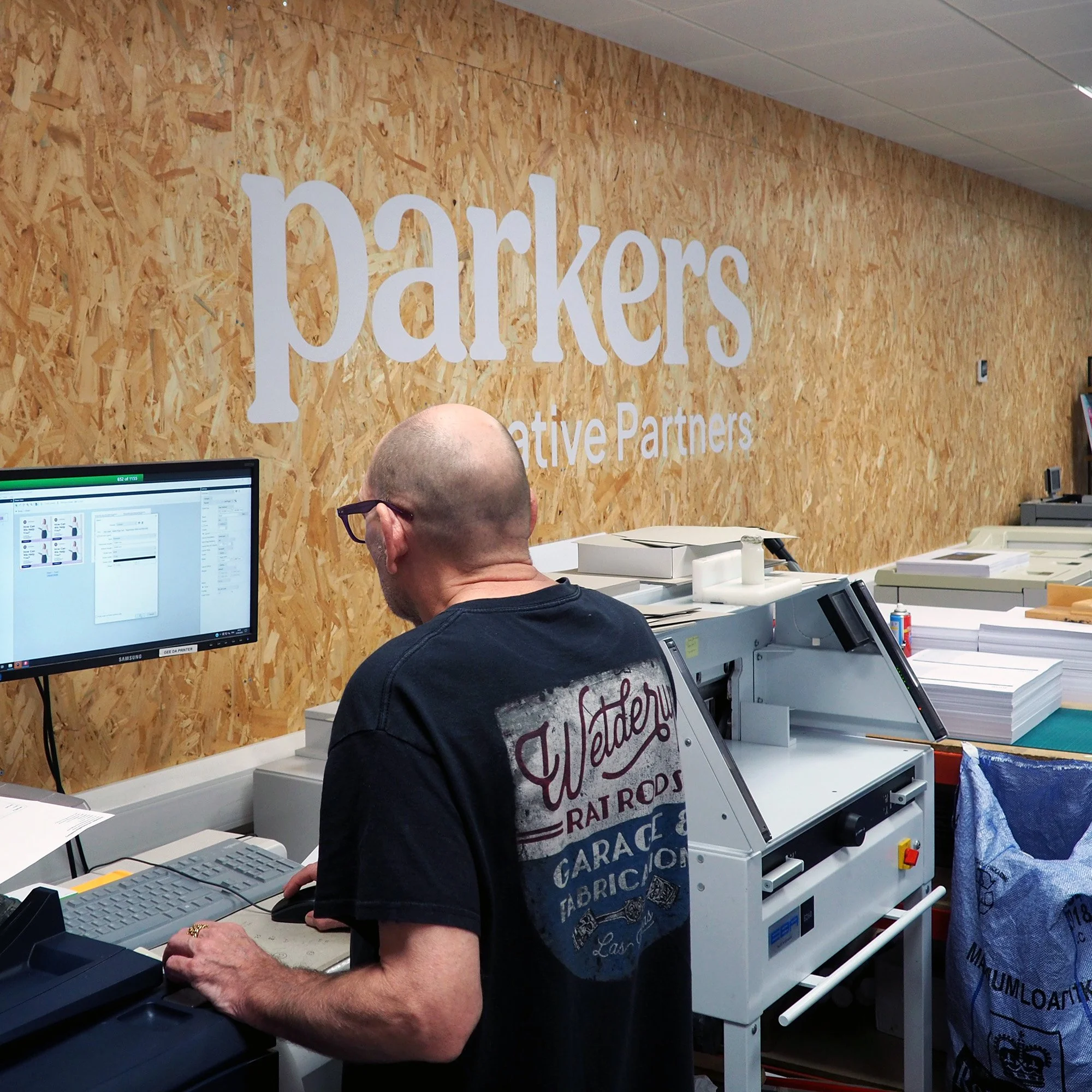 A person with a bald head and glasses standing at a workstation with a computer and printing equipment, inside an office with a wood-textured wall that has 'Parkers Creative Partners' written on it.