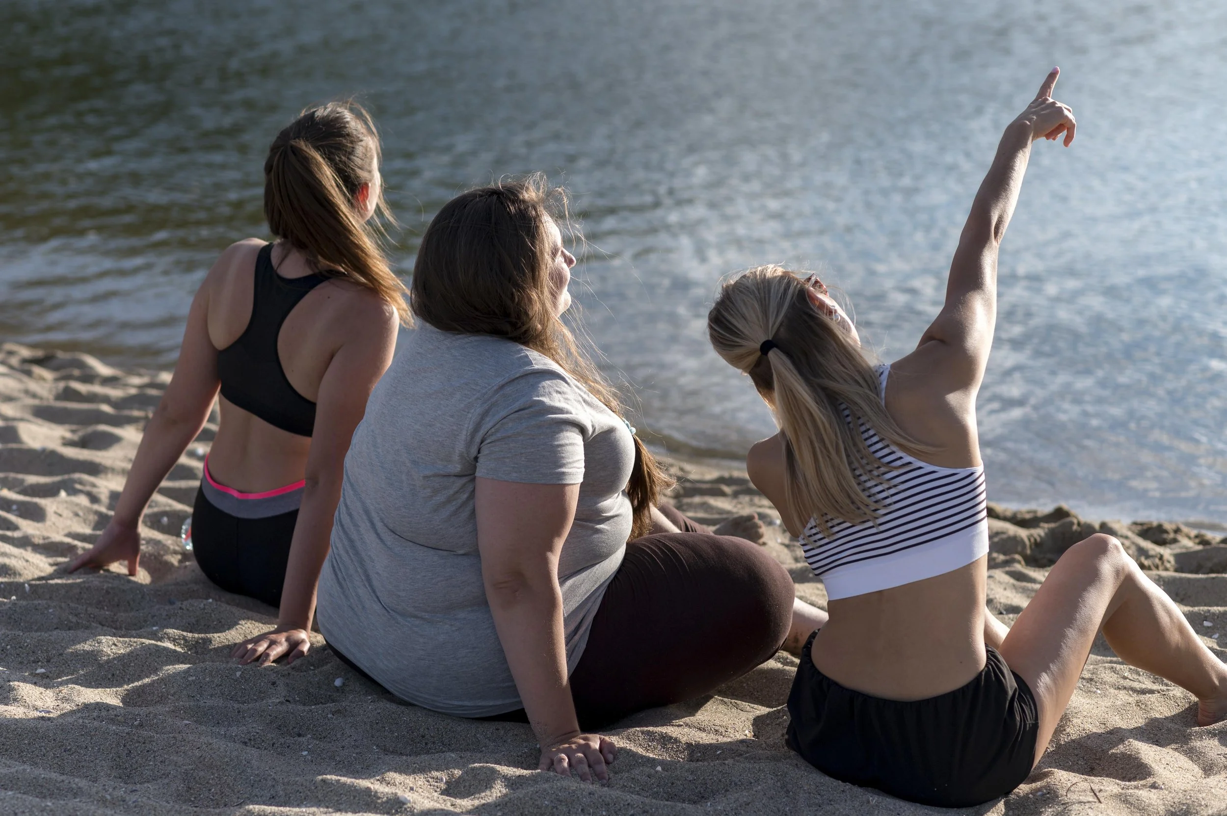 Three women sitting on a sandy beach by the water, one pointing towards the water.