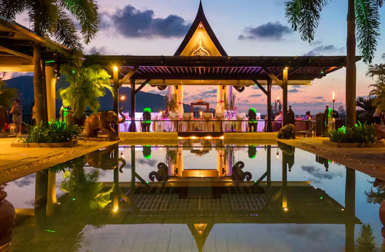 A scenic view of an outdoor pavilion with Thai-style architecture at sunset, reflected in a calm pool, decorated with plants, elephants sculptures, and colorful lighting.