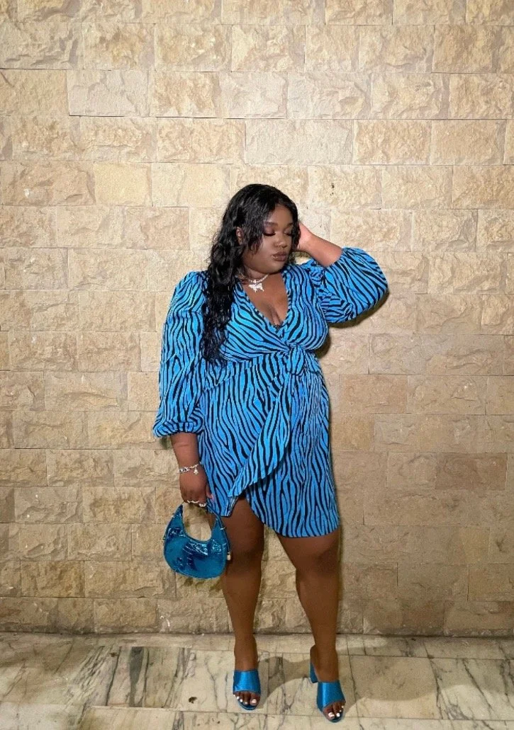 Woman with long black wavy hair wearing a blue zebra print dress, matching blue high heels, and blue purse, standing against a beige stone wall with her hand resting on her head.