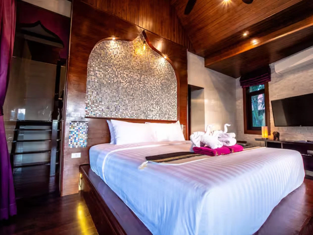 A bedroom with a bed decorated with towel art resembling swans, in front of a wooden headboard with mosaic tile accents, a flat-screen TV, window with purple curtains, and warm lighting.