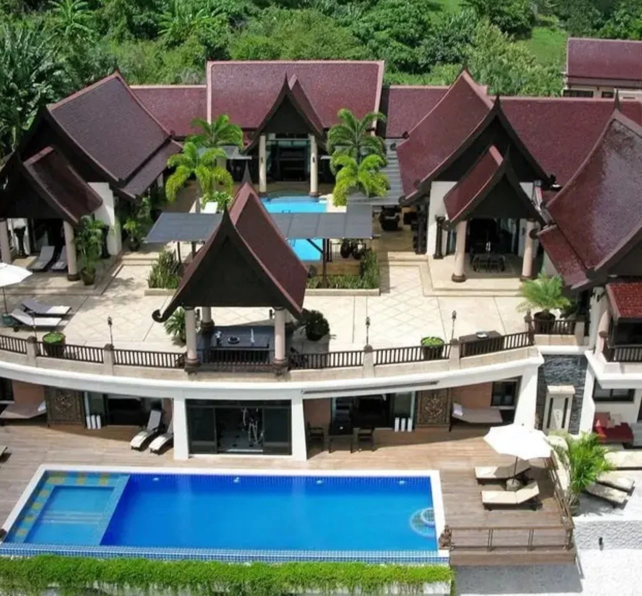 Aerial view of a luxurious beachfront villa with a swimming pool, landscaped gardens, and multiple traditional style rooftops surrounded by lush green trees.