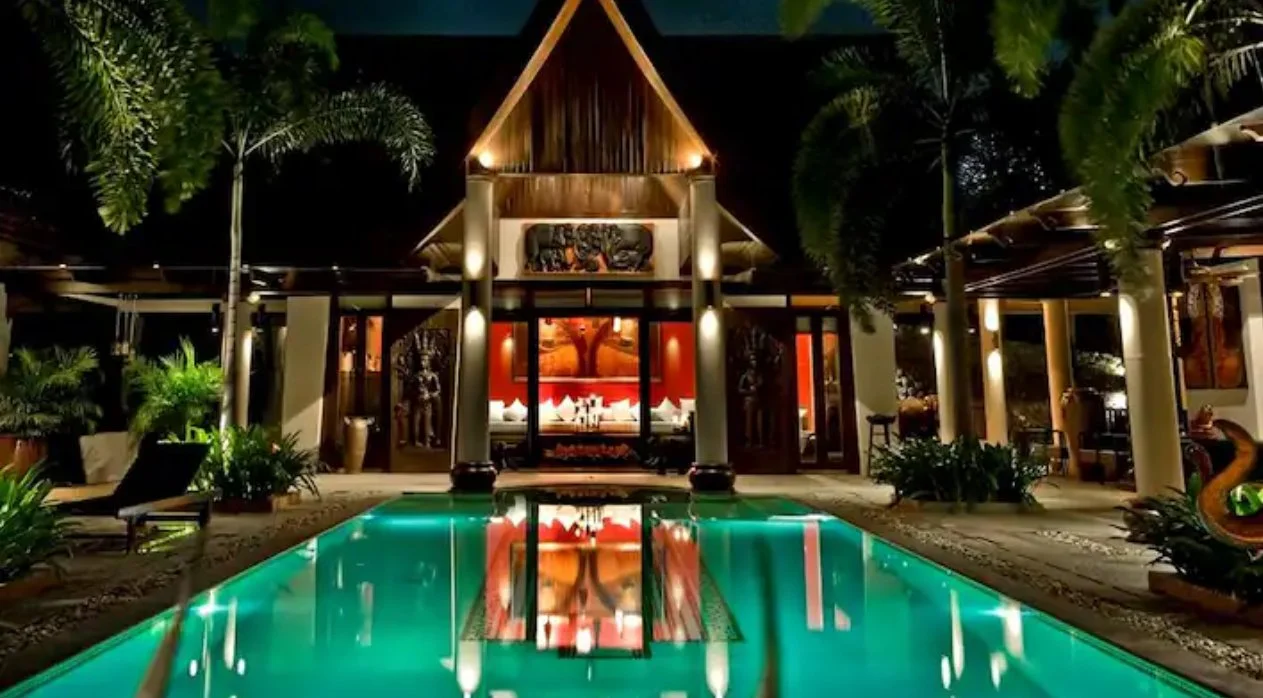 Luxurious tropical resort with a swimming pool at night, illuminated by surrounding lights, with traditional wooden architecture and lush greenery.