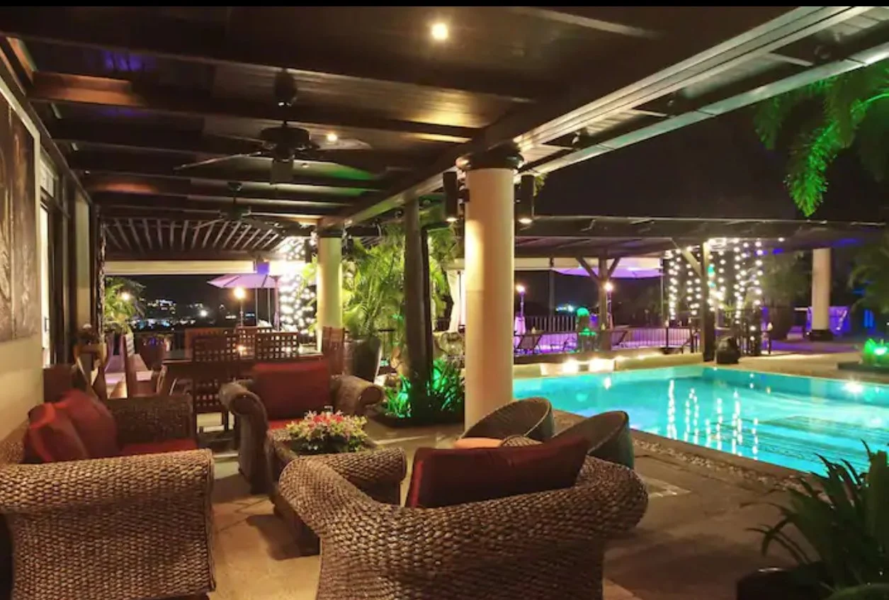 Nighttime outdoor pool area with wicker lounge chairs and seating, illuminated by green and purple lighting, with string lights wrapped around trees and umbrellas providing ambiance.