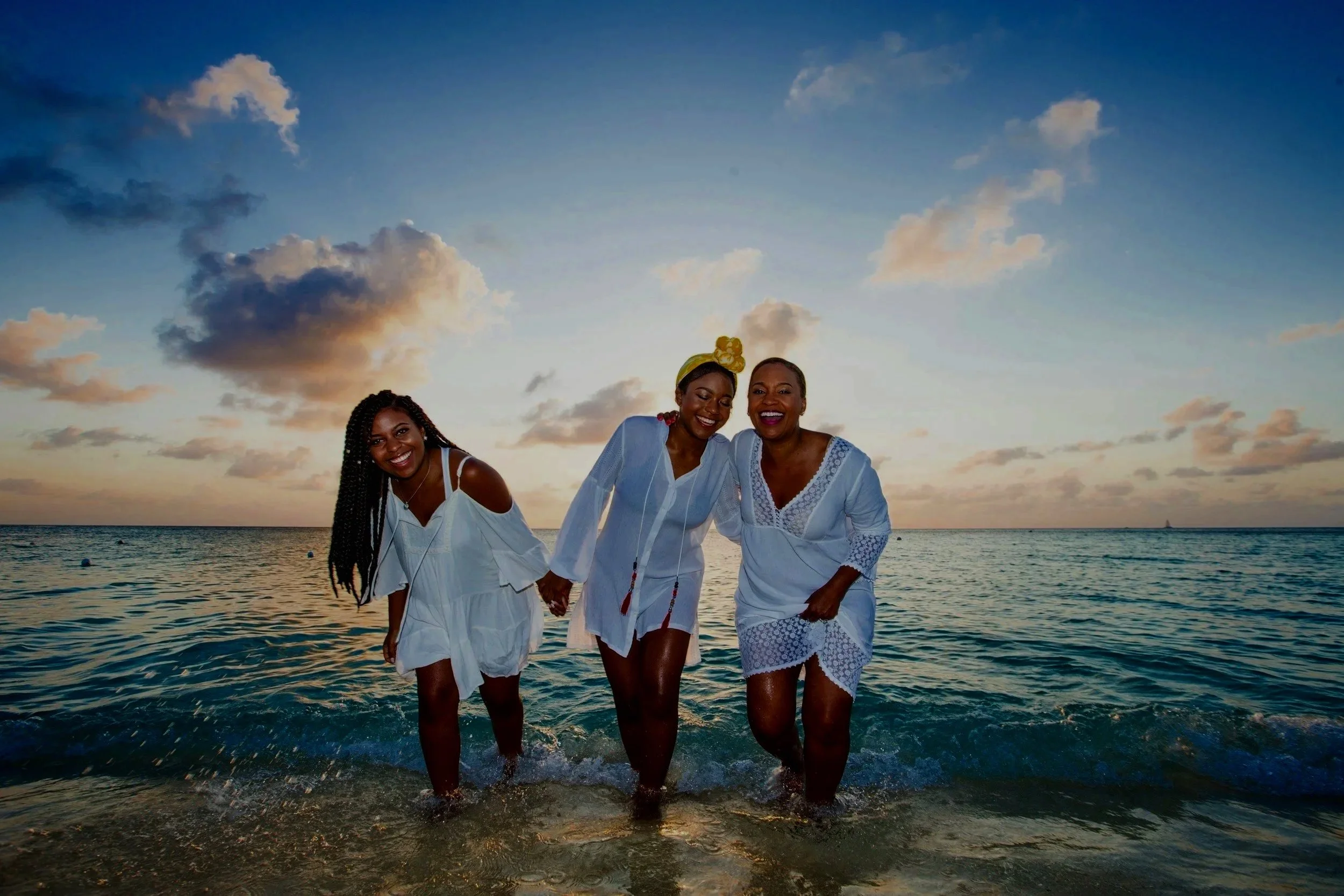 Three women in white dresses walking and holding hands in the ocean at sunset, smiling and enjoying the beach.