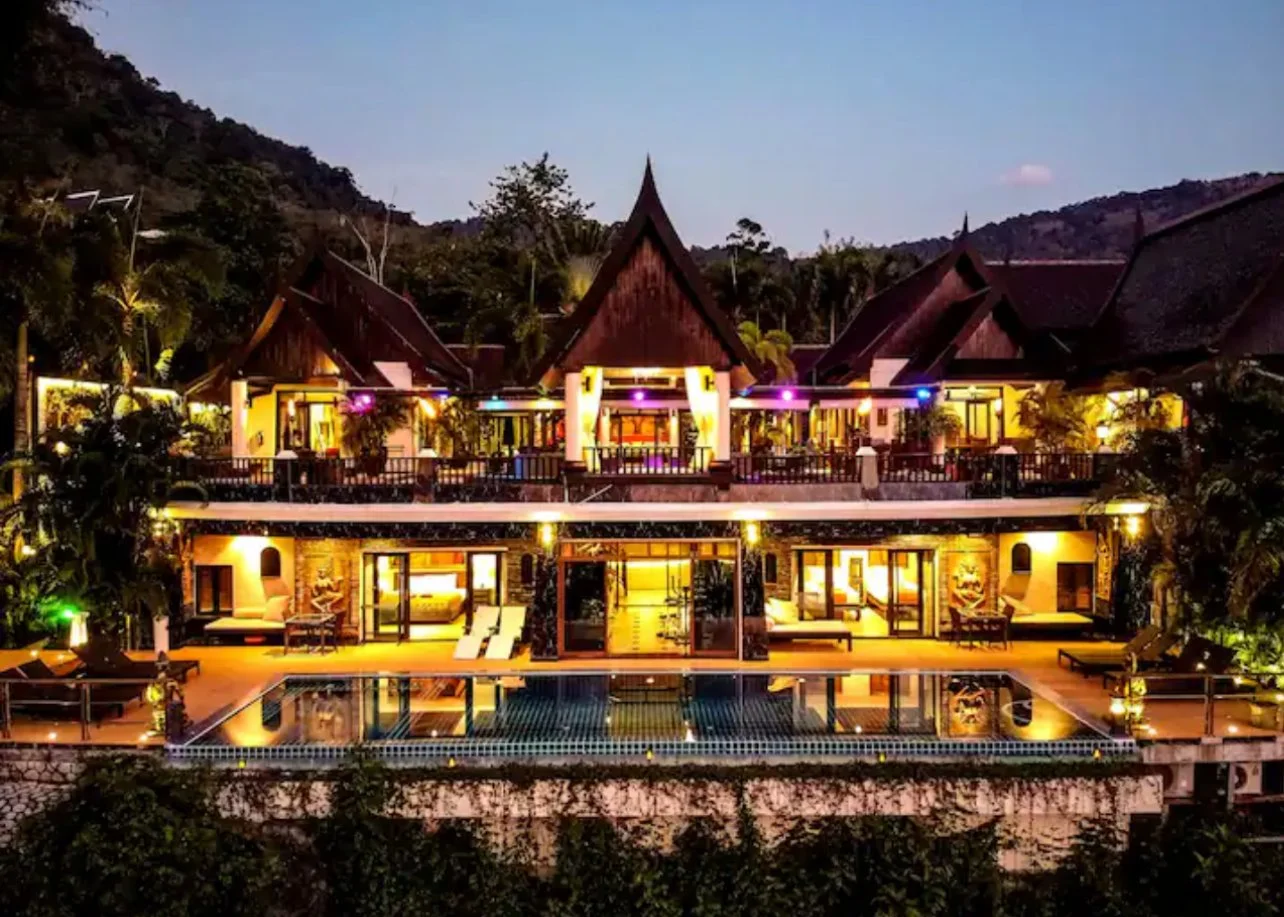 Luxury multi-story resort illuminated at night with traditional Thai-style architecture, surrounded by trees and mountainous landscape.