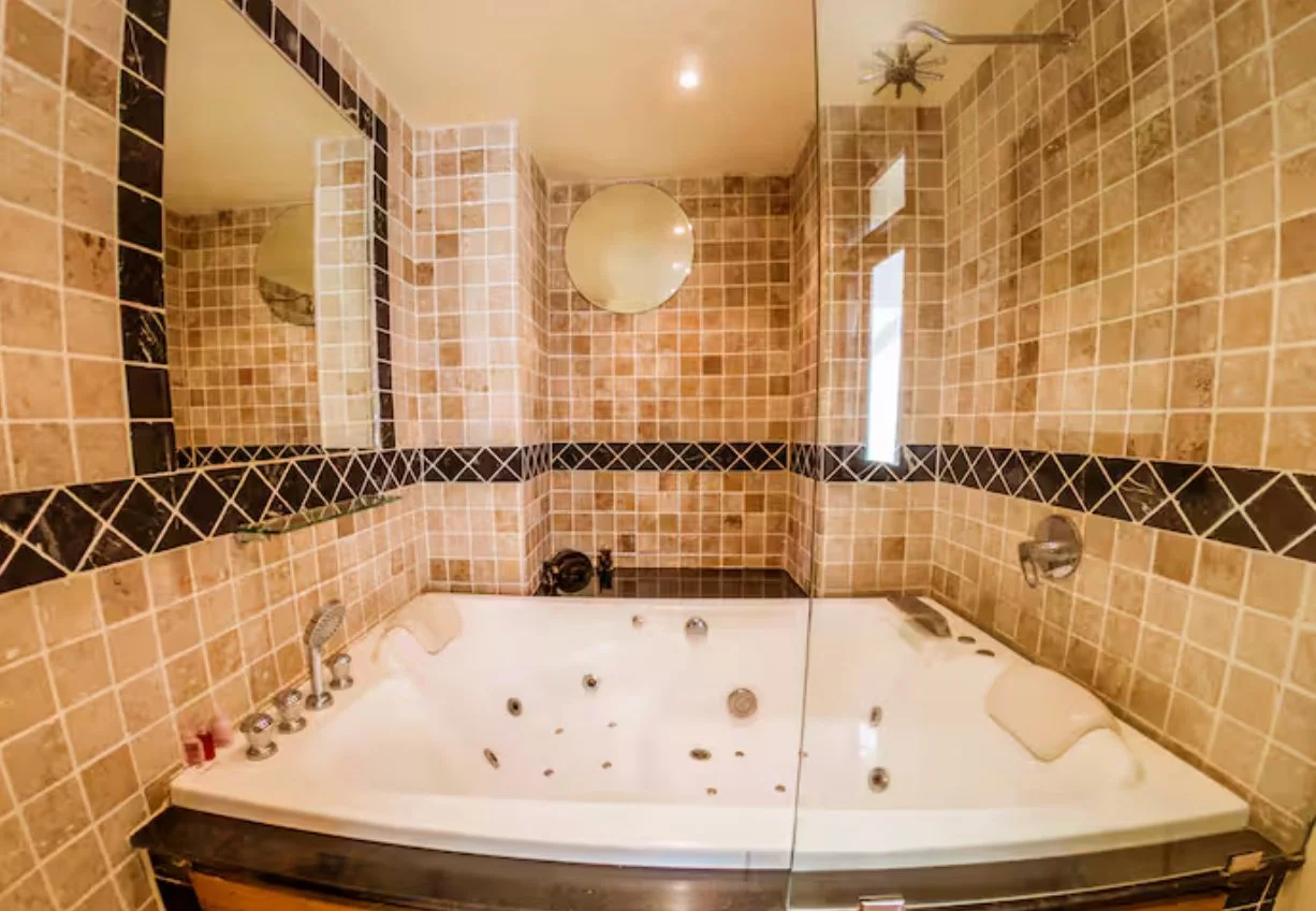 A bathroom with tiled walls and a large bathtub with jets, mirrors, and a glass partition. The tiles are beige with black border accents. There is a ceiling showerhead and a window providing natural light.