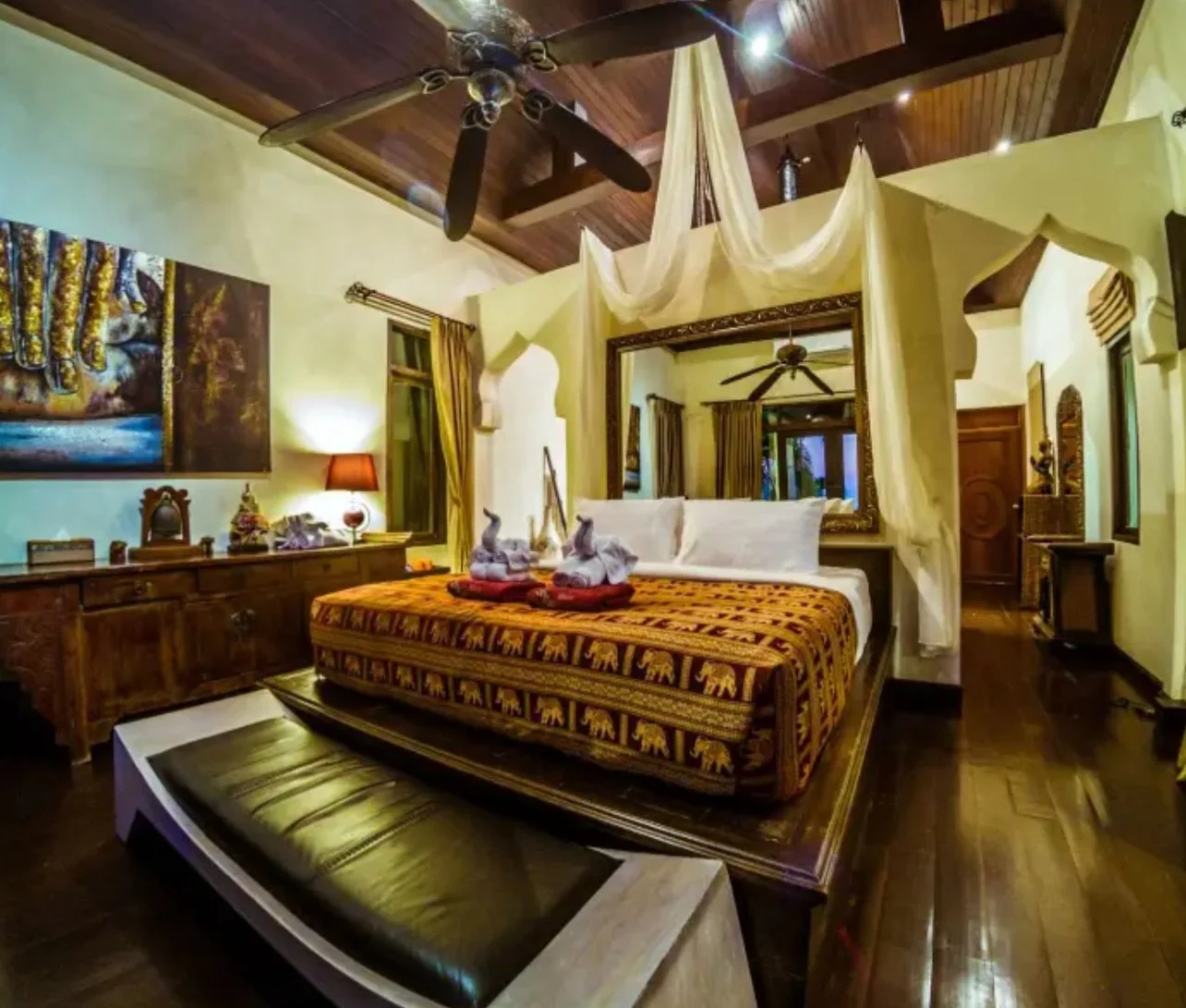 Luxurious bedroom with a four-poster bed, wooden furniture, and tropical decor, featuring a ceiling fan, swan towel sculptures on the bed, and artwork on the wall.