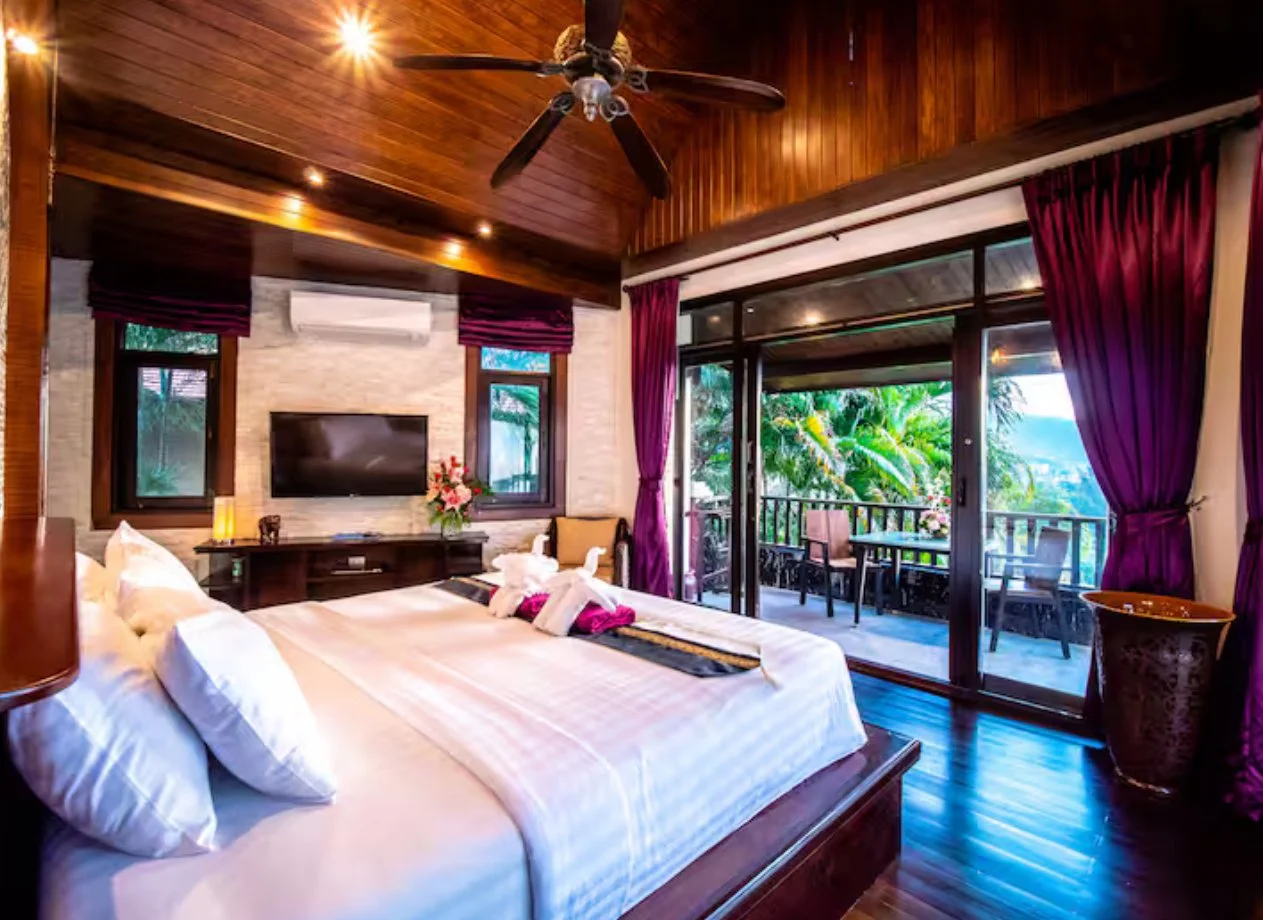 A luxurious bedroom with a wooden ceiling, large bed with white linens and towels folded as swans, a flat-screen TV, a small balcony outside with chairs, and tropical greenery visible through glass sliding doors.