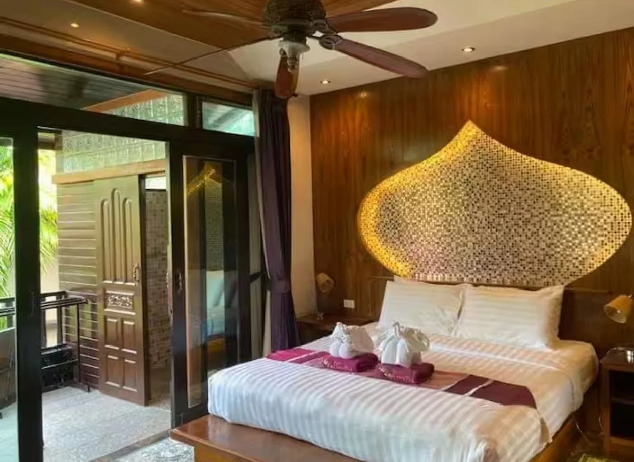 Hotel room with a wooden wall, decorative headboard above the bed, ceiling fan, sliding glass door leading to a balcony with tropical plants.