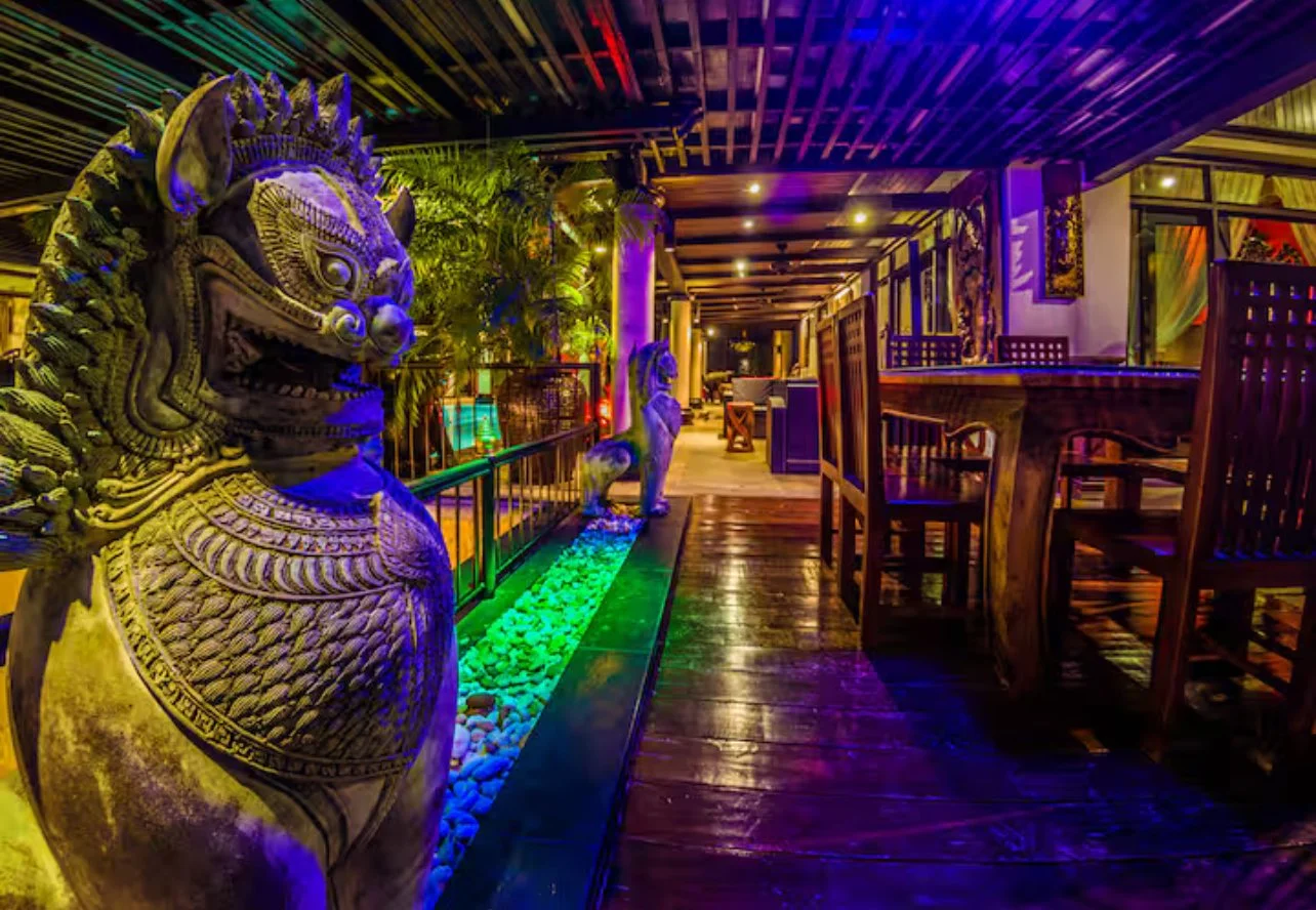 Colorfully lit outdoor restaurant or bar with wooden furniture and Asian-inspired sculptures, including a large lion statue, under a wooden roof.