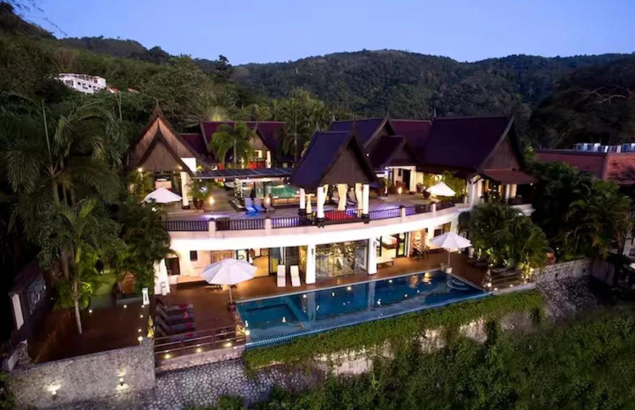 A luxurious villa with a swimming pool, outdoor seating, and tropical landscaping, set against a backdrop of green hills at dusk.
