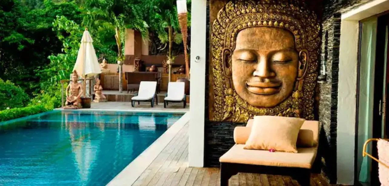 Luxury outdoor pool area with lounge chairs, shaded umbrellas, tropical greenery, and a large carved Buddha face wall sculpture with a small covered seating area.