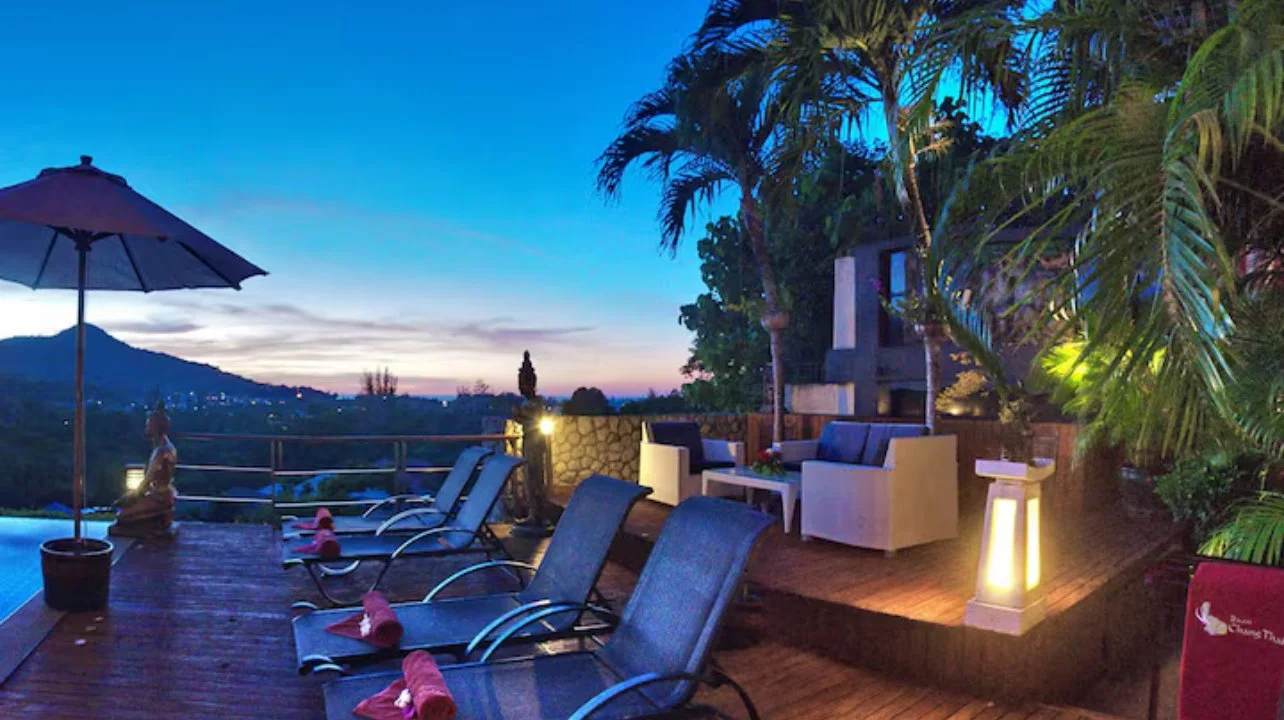 An outdoor poolside area at dusk with lounge chairs, an umbrella, and a seating area with couches, surrounded by palm trees and landscape lighting.