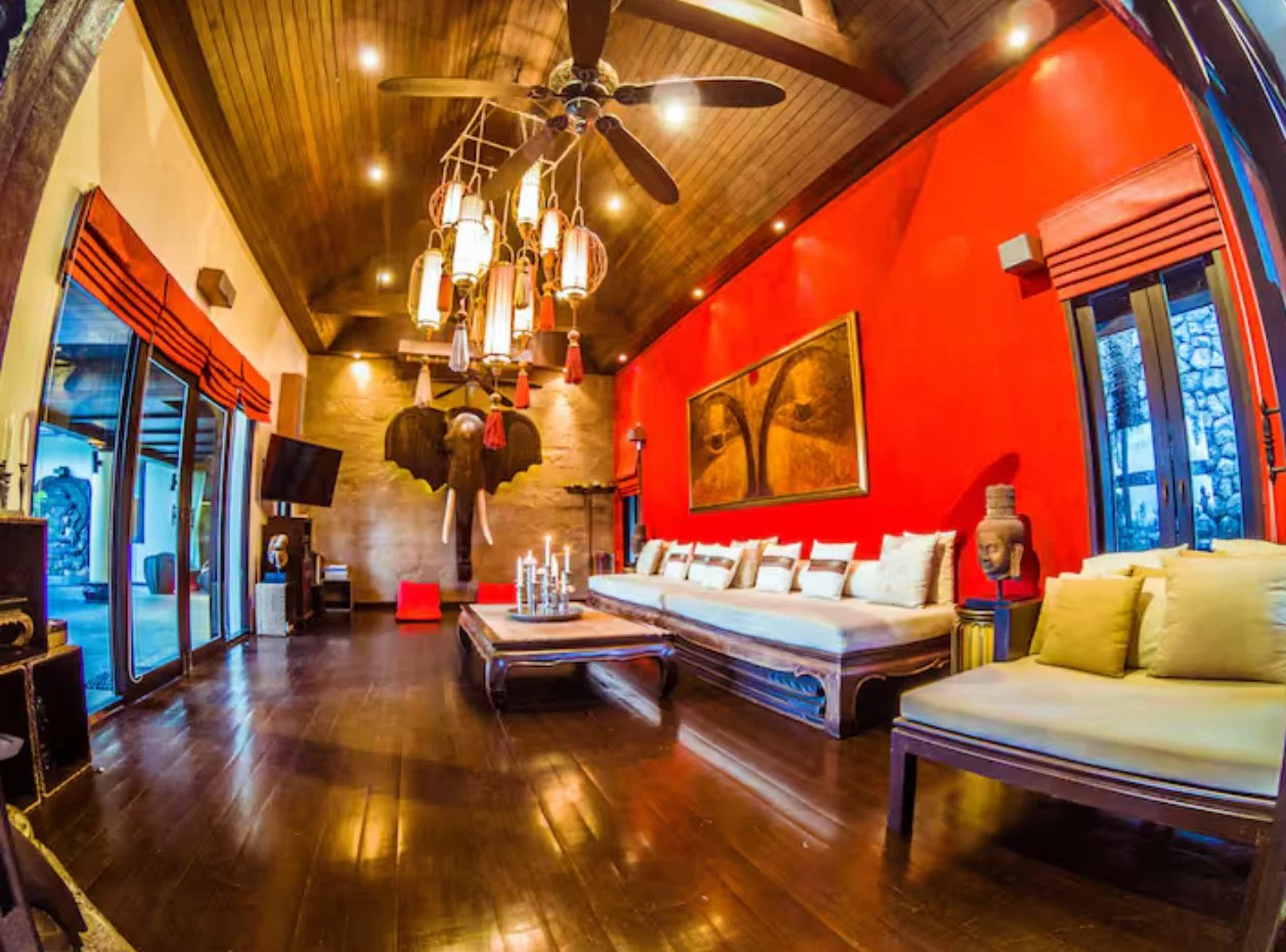 Living room with red accent wall, wooden furniture, decorative art, and colorful lighting fixtures hanging from a wooden ceiling.