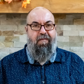 A man with glasses and a full beard, wearing a dark blue, dotted button-up shirt, standing in front of a wall with brick and wood paneling.