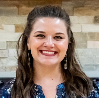 A woman with long, wavy hair smiling in front of a stone wall background.