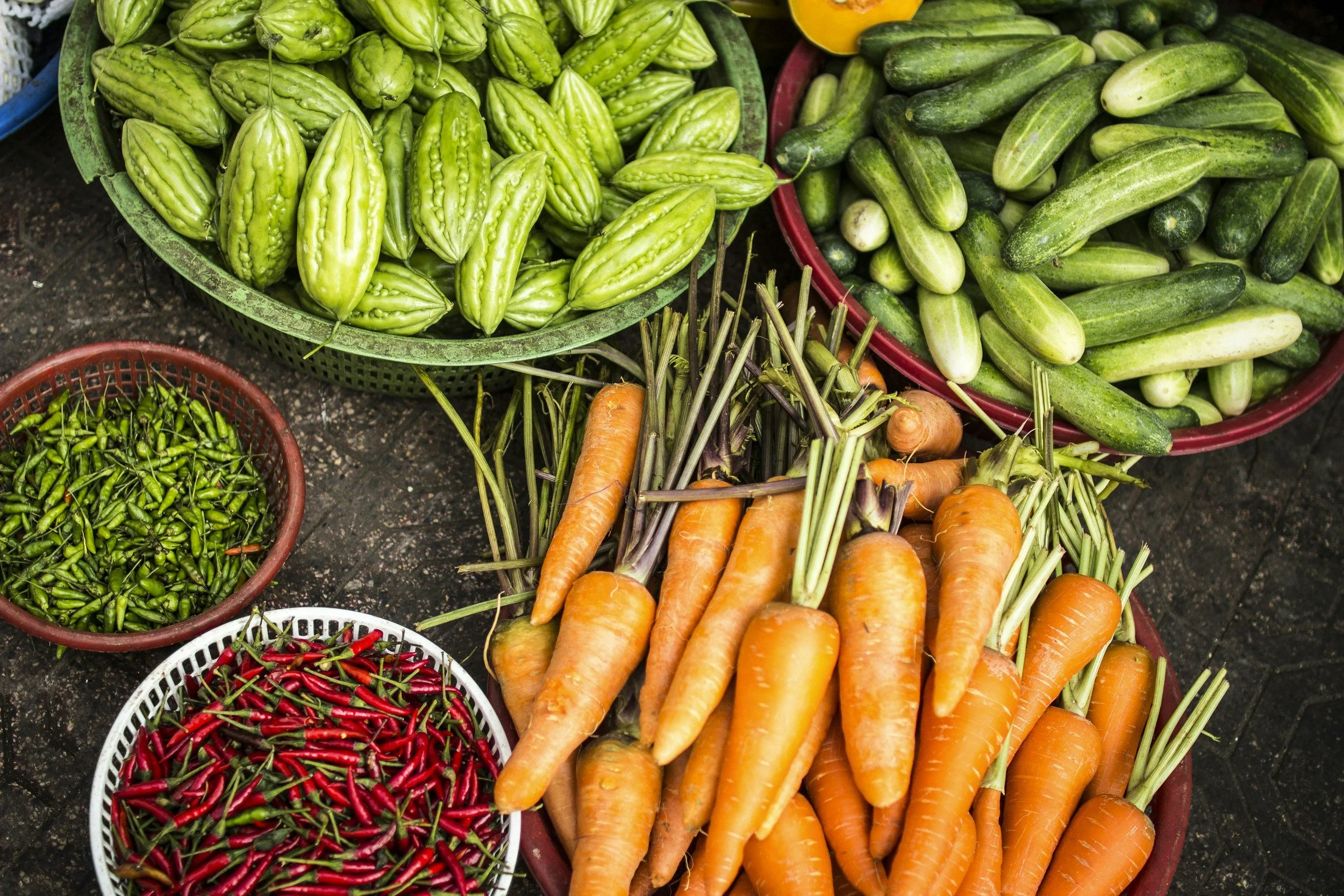 Basket of green bitter melons, basket of large cucumbers, basket of red and green chili peppers, and bunches of orange carrots.