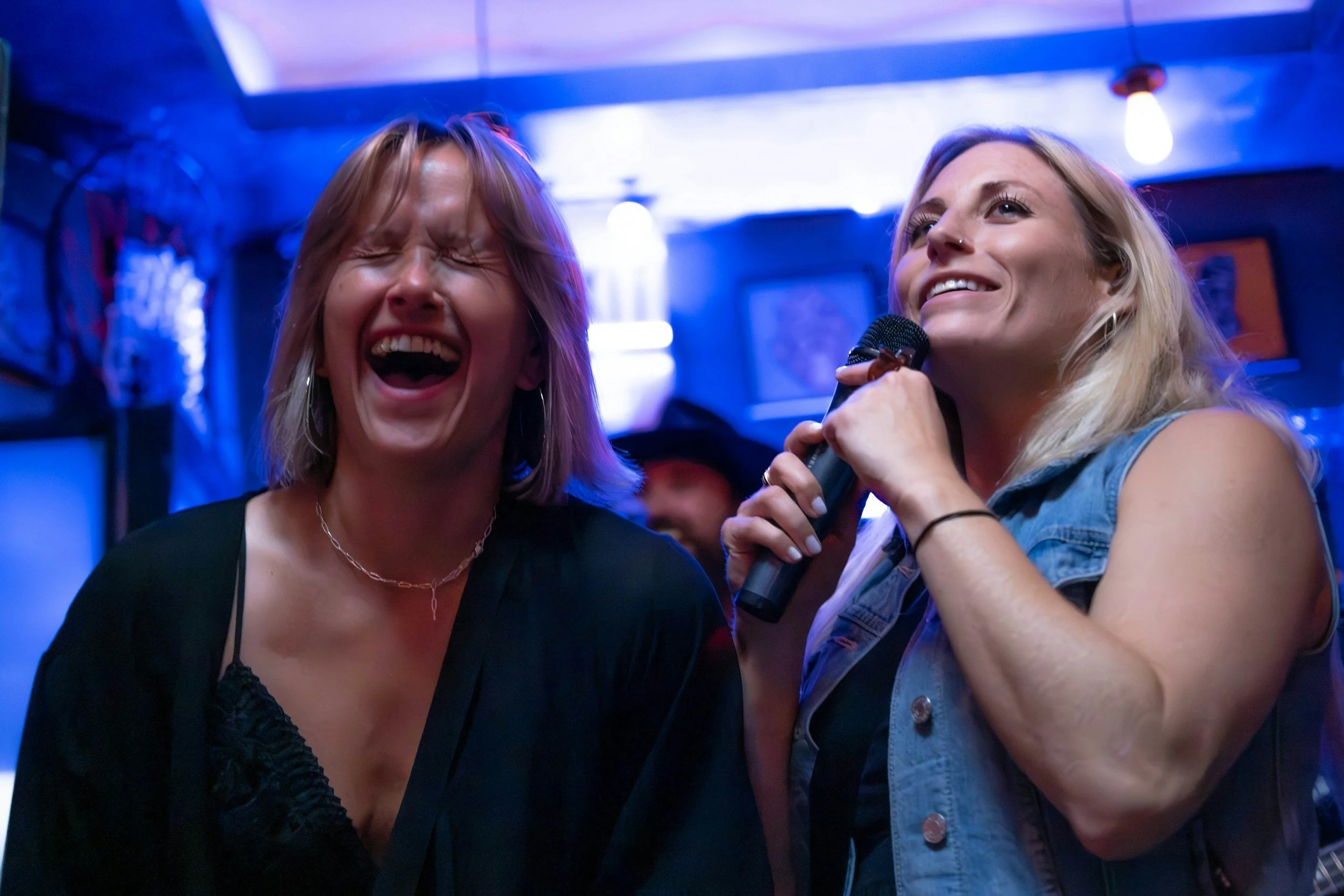 Two women singing and laughing in a dimly lit bar or club. One woman has blonde hair and is holding a microphone, smiling, while the other woman has light brown hair and is laughing with her eyes closed.